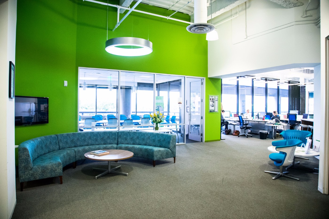 Modern open-plan office lounge with curved teal sofa, round coffee table, and bright green accent wall