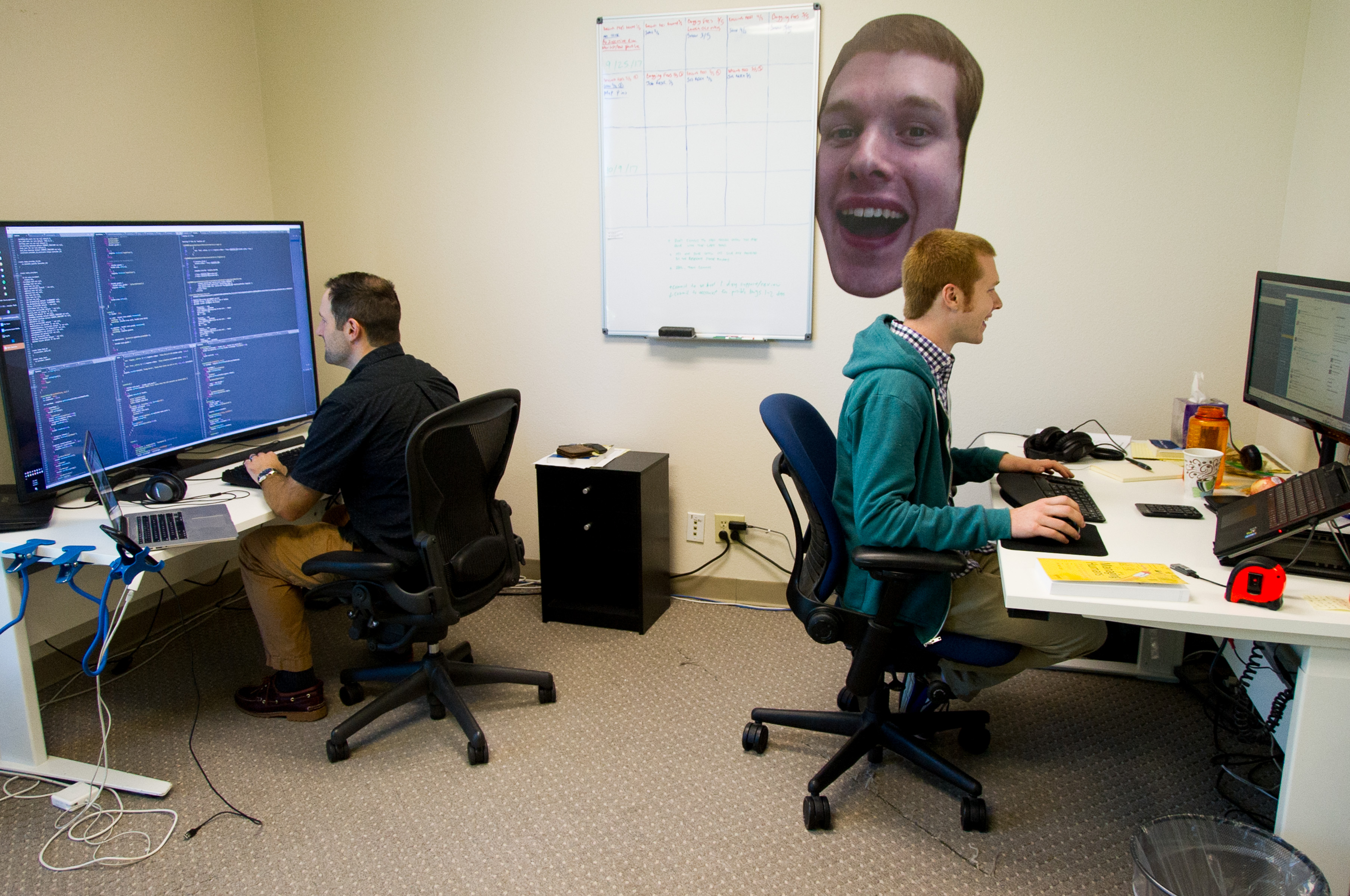 Two Lawn Love engineers coding at desks with giant cardboard cutout face poster of colleague hanging on the wall