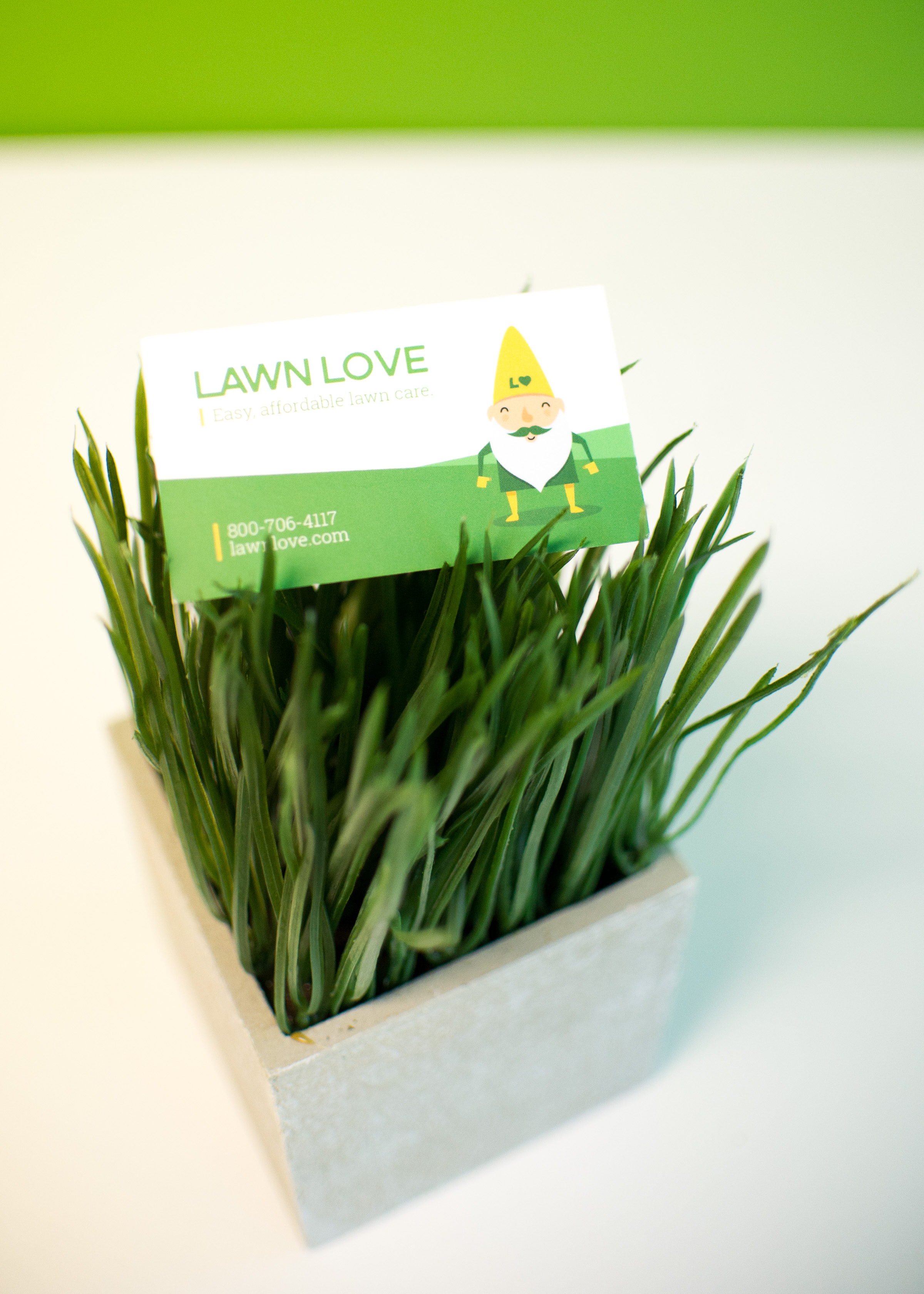 Lawn Love business card with gnome mascot logo displayed in small grass-filled planter on white desk