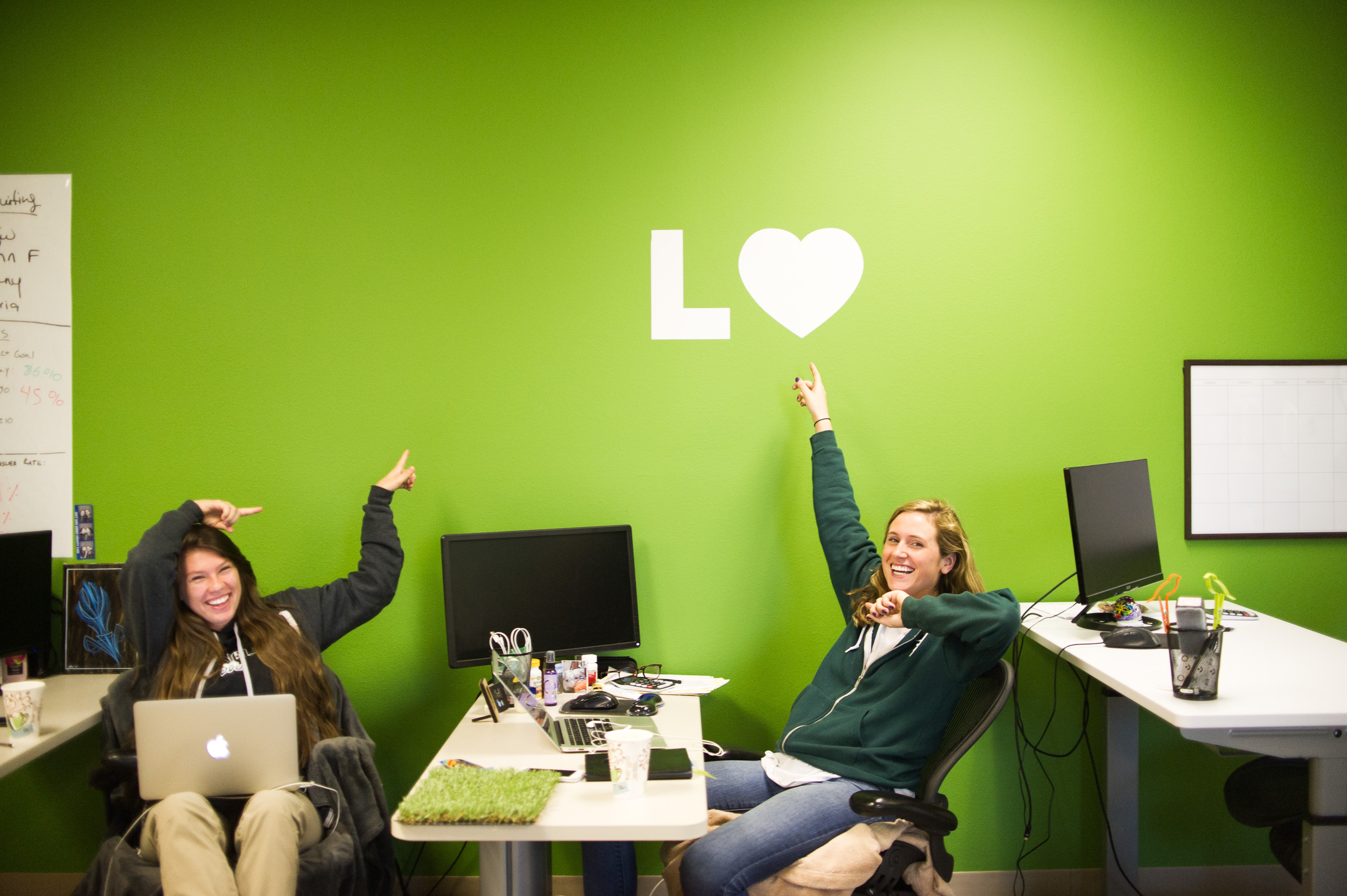 Two Lawn Love team members laughing while pointing up at the L heart Lawn Love logo on green office wall