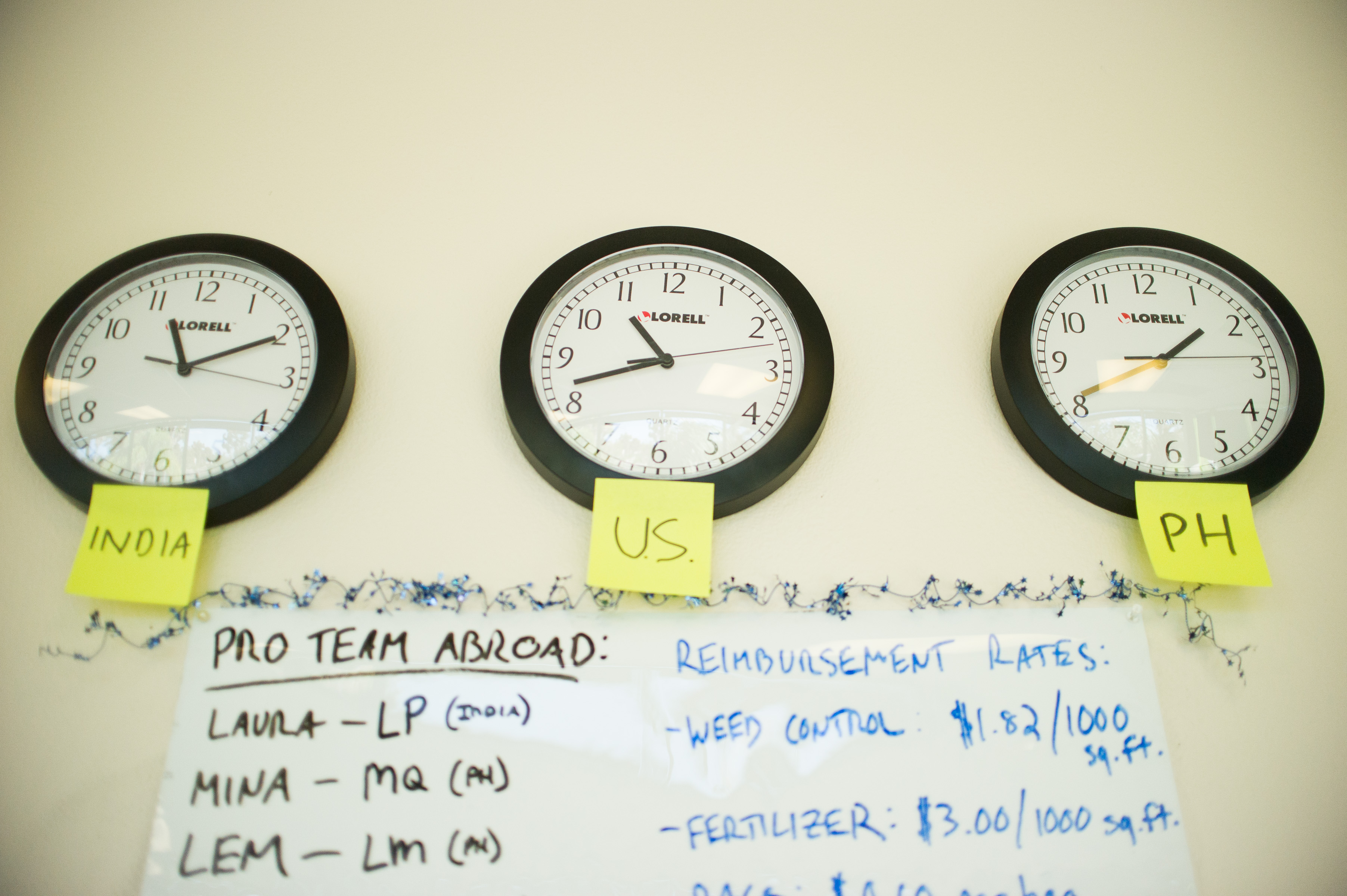 Three wall clocks at Lawn Love office showing time zones for India, U.S., and Philippines with whiteboard listing remote pro team