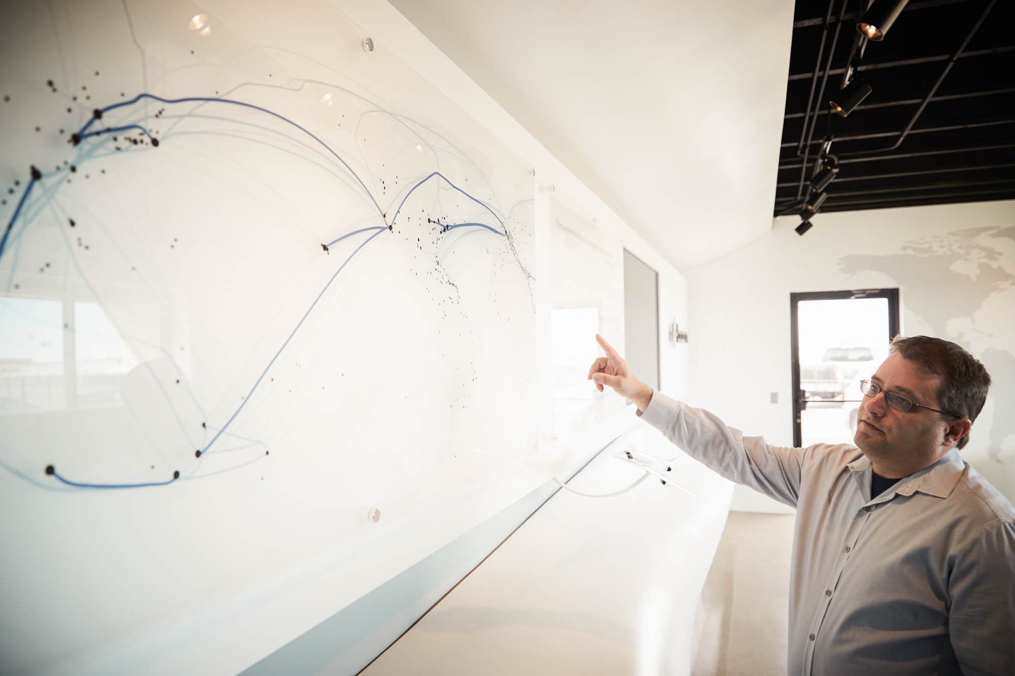 Boom Supersonic team member pointing at wall-mounted glass display showing global flight route map