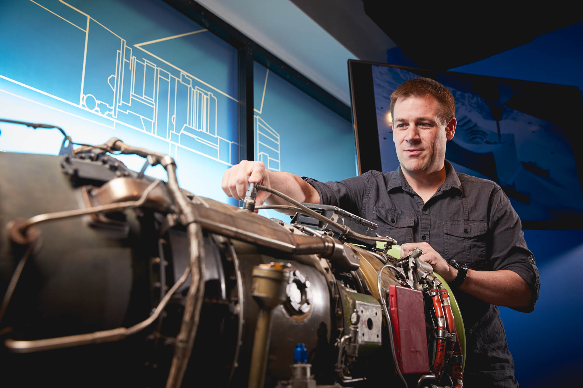 Boom Supersonic engineer in a dark shirt working on a disassembled jet engine with engineering schematics displayed on illuminated screens behind