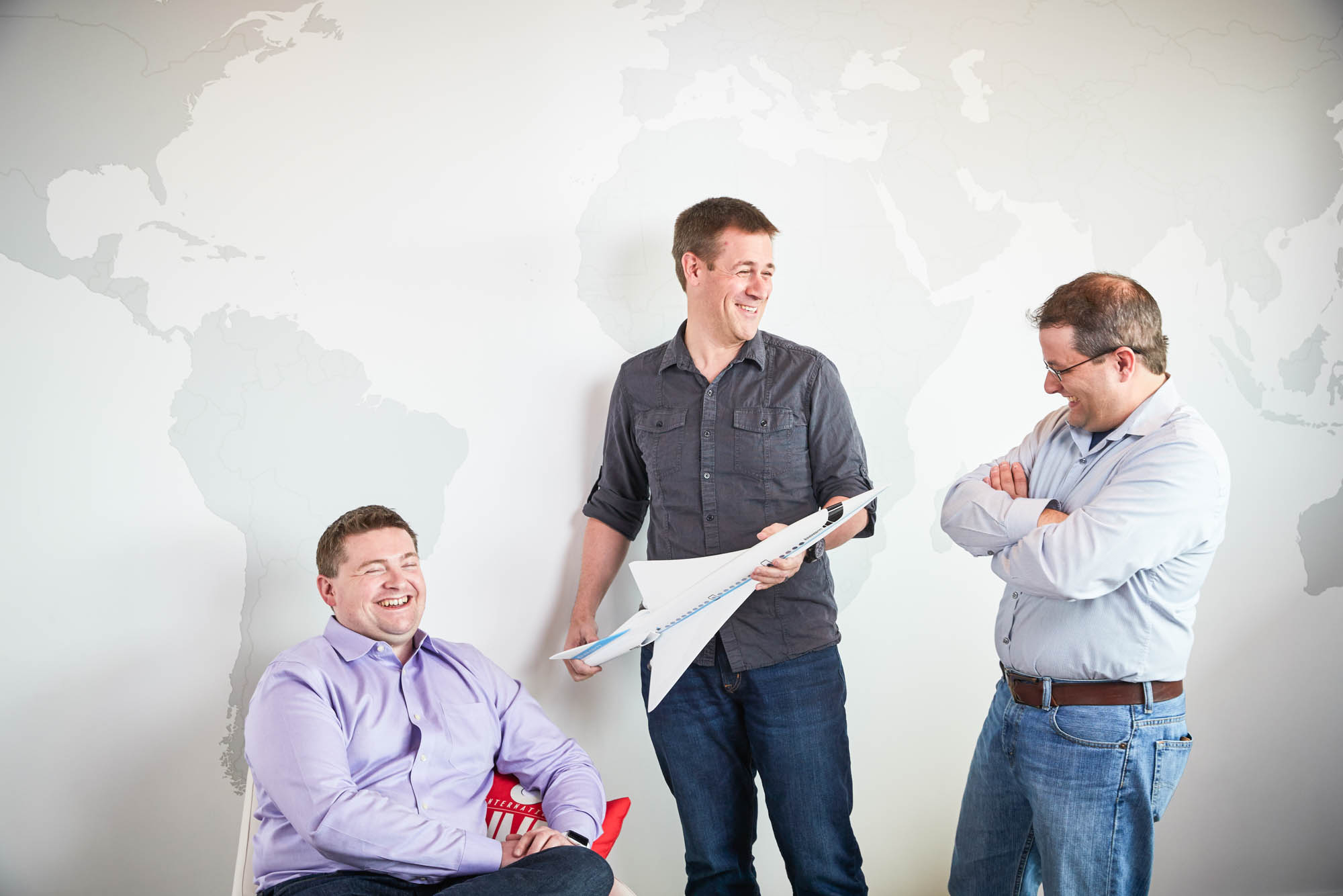 Three Boom Supersonic founders laughing and discussing while holding a paper airplane model in front of a world map wall