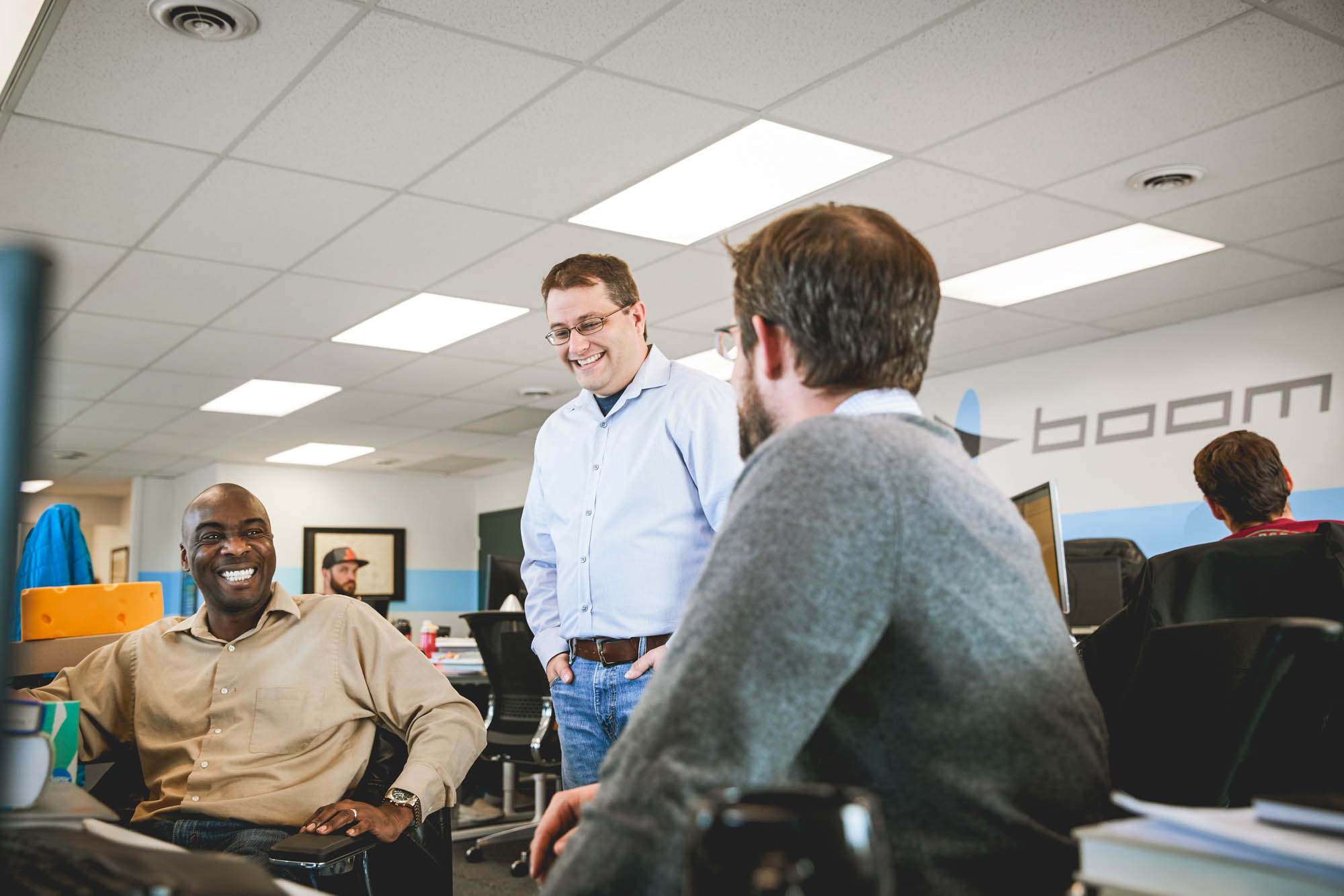 Three Boom Supersonic team members chatting and laughing in their open-plan office with the Boom logo painted on the wall behind them