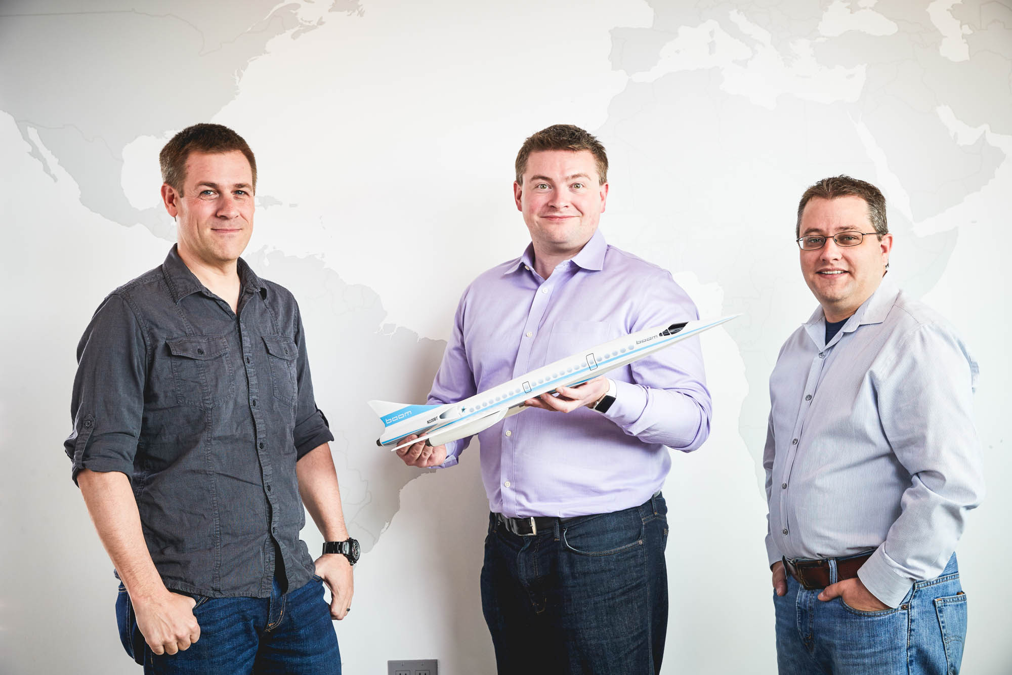 Three Boom Supersonic founders posing together with a model of their supersonic airliner against a wall mural of the world map