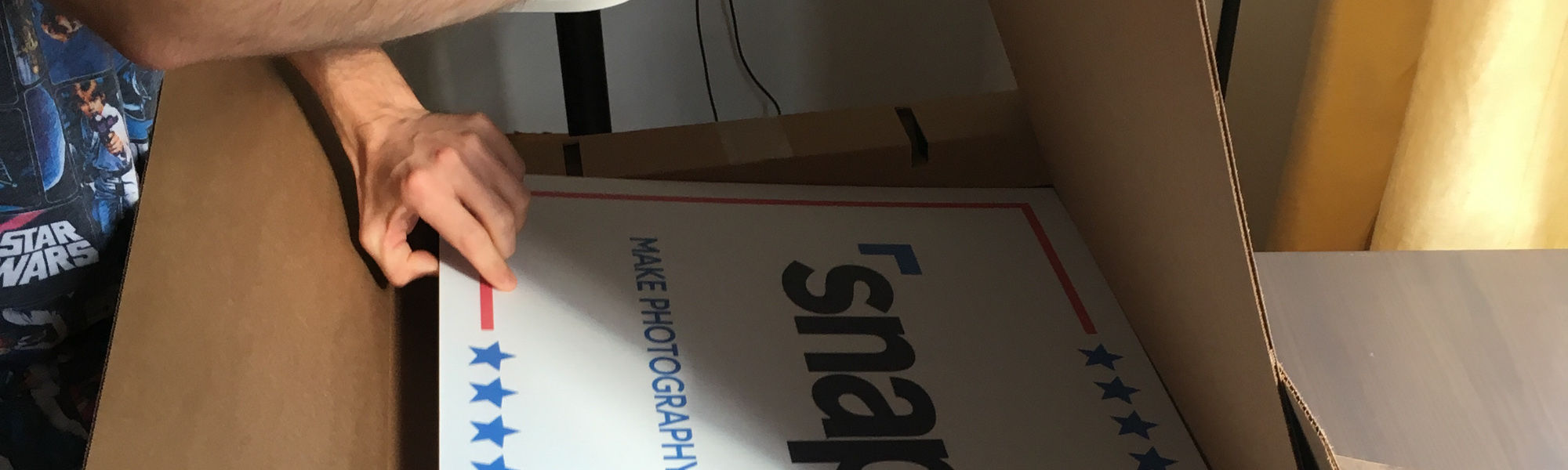 Hand pulling Snappr Make Photography Great Again campaign poster from a moving box