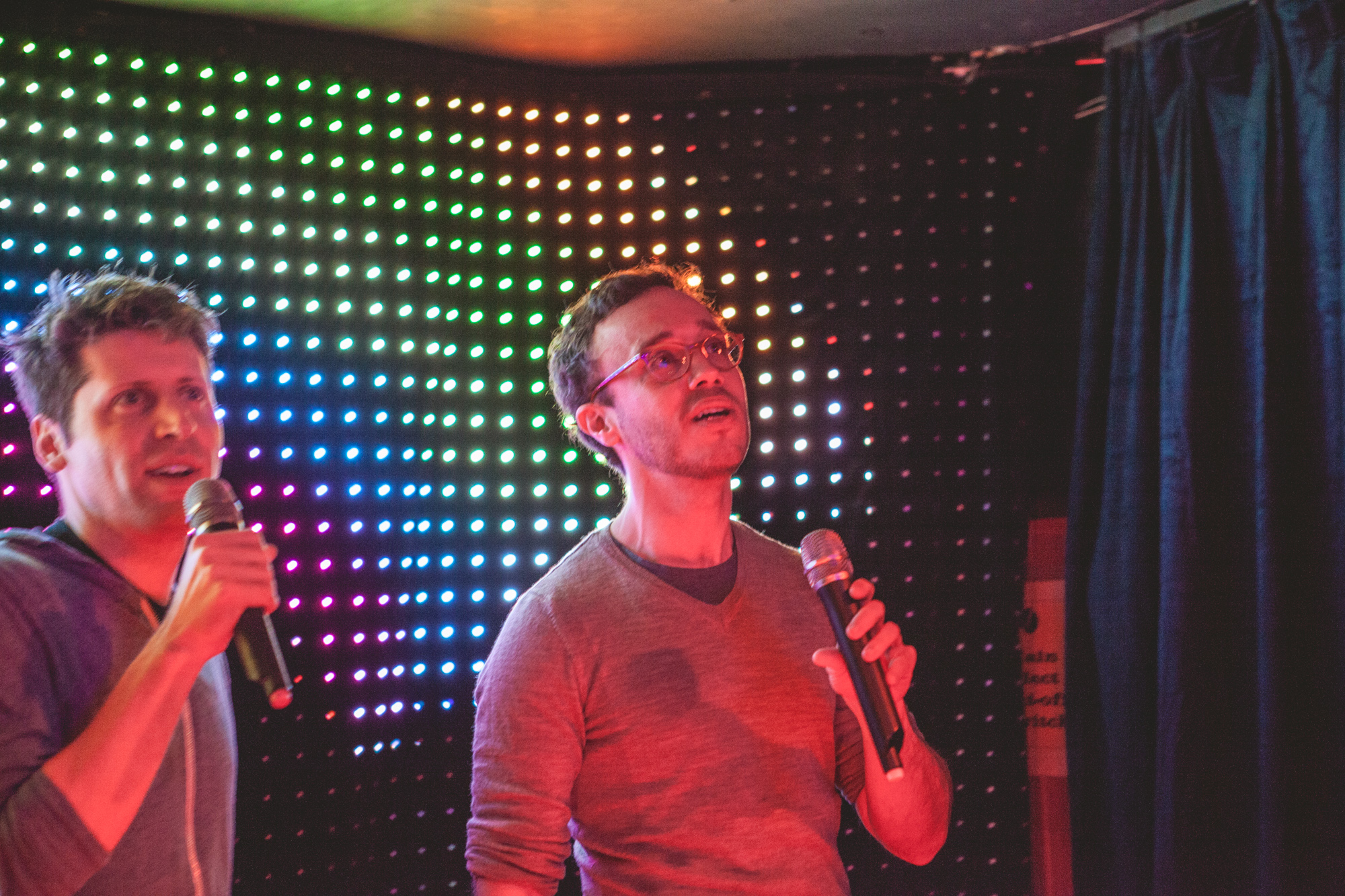 Two Snappr cofounders singing karaoke with microphones in front of colorful LED light backdrop