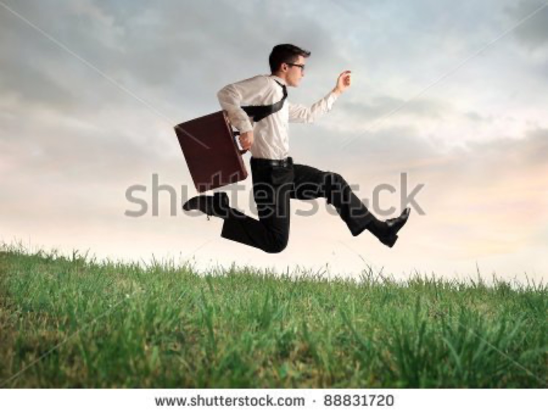 Businessman in a tie leaping mid-air over a green meadow while holding a briefcase, a classic overused businessman-in-a-field stock photo cliche
