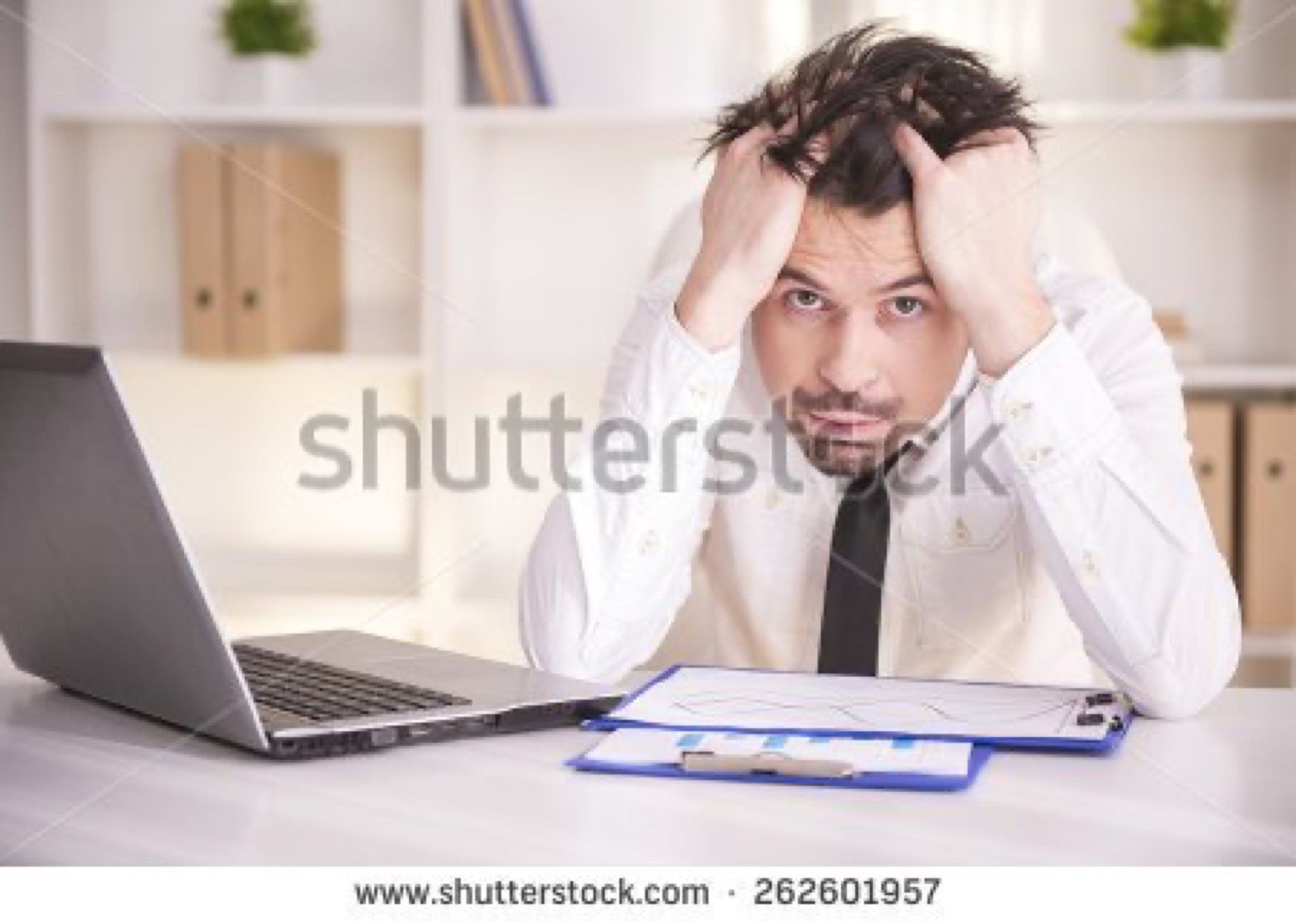 Frustrated man in a white shirt gripping his head at a laptop in an office, one of the most overused stressed technology guy stock photo cliches