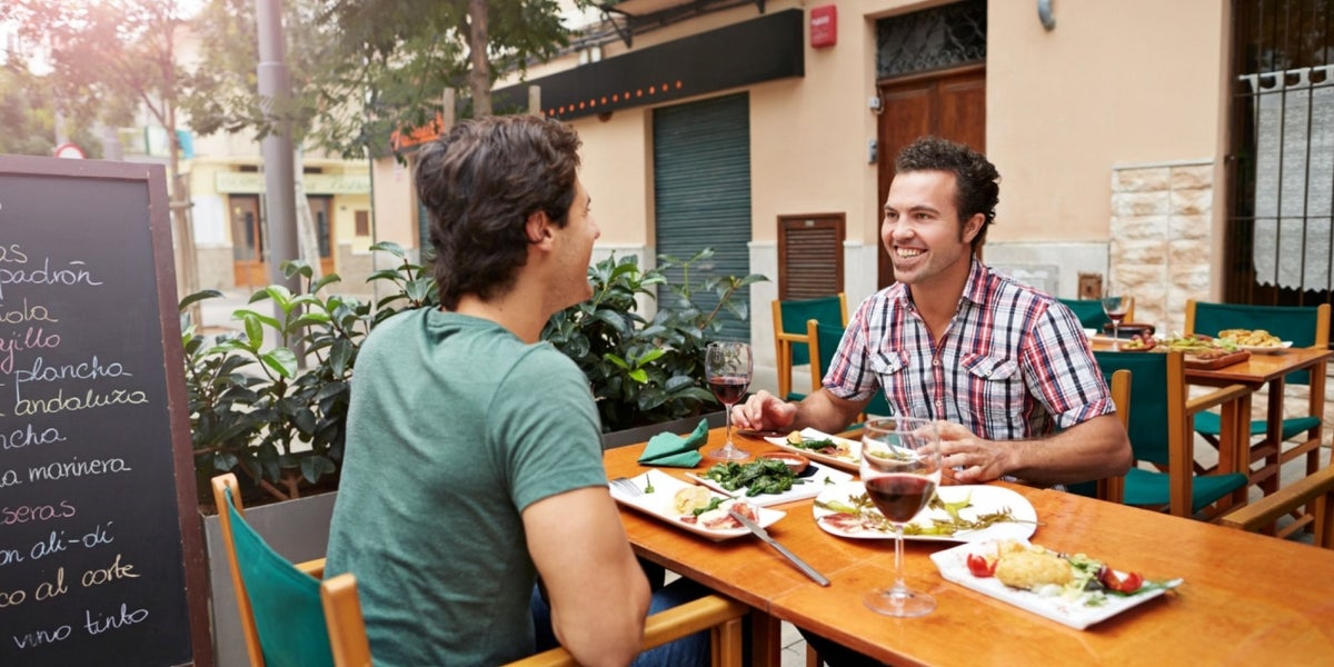 Two diners laughing over food and wine at a sunlit outdoor café terrace