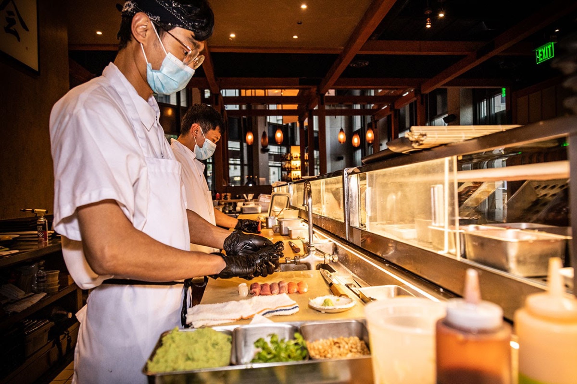 Masked sushi chefs preparing nigiri at a Japanese restaurant counter with warm ambient lighting