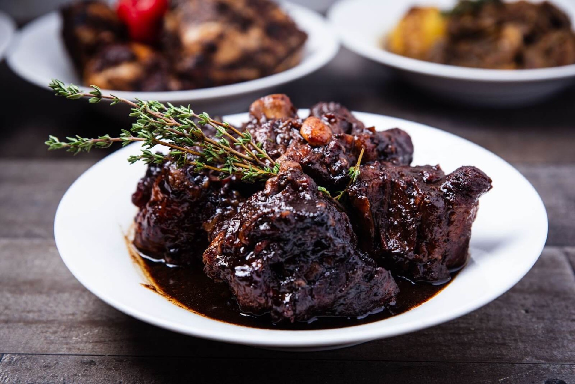 Braised oxtail in dark sauce with fresh thyme on a white plate, illustrating quality food imagery for delivery apps