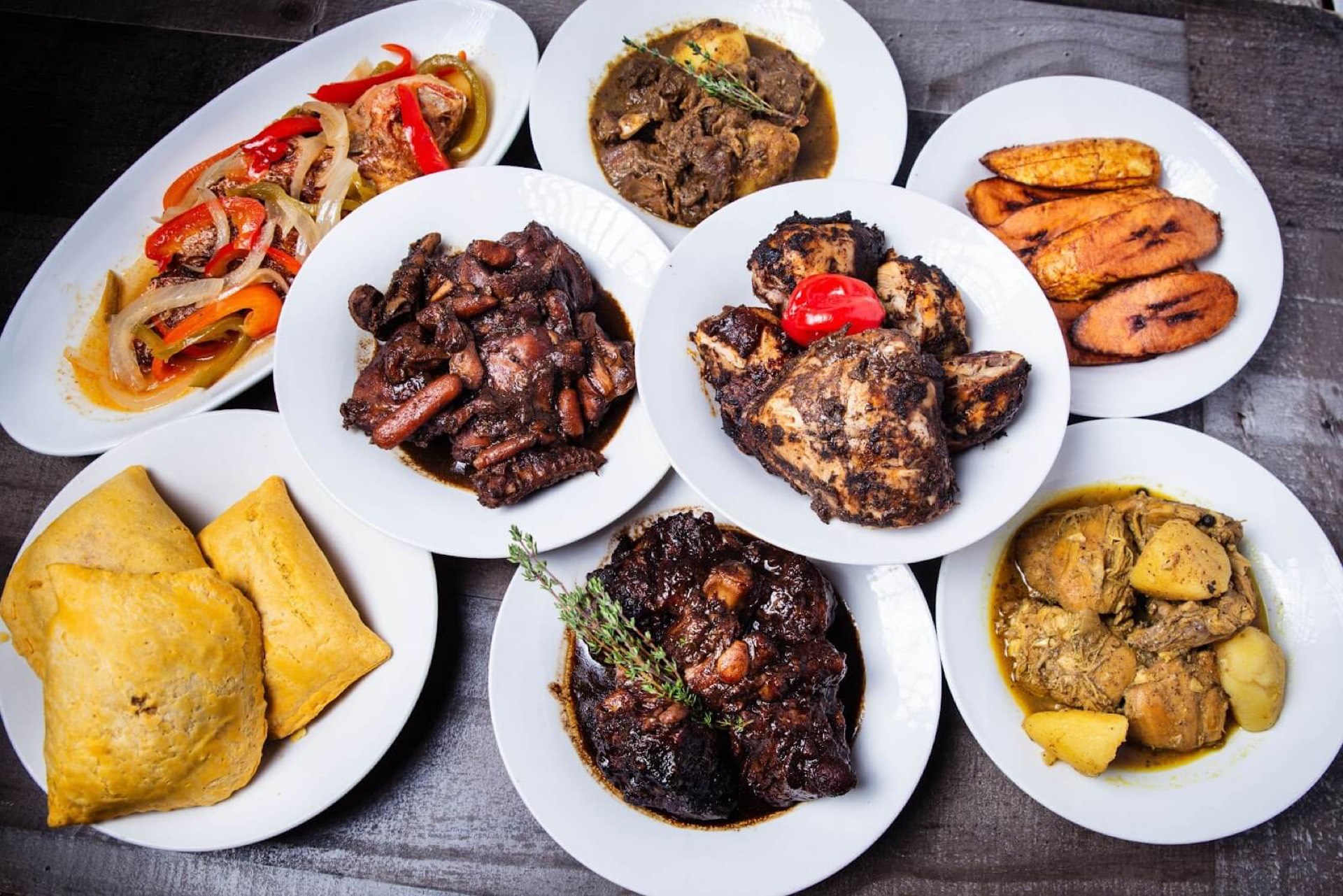 Overhead spread of eight Caribbean dishes including jerk chicken, oxtail, plantains and patties for a food delivery menu