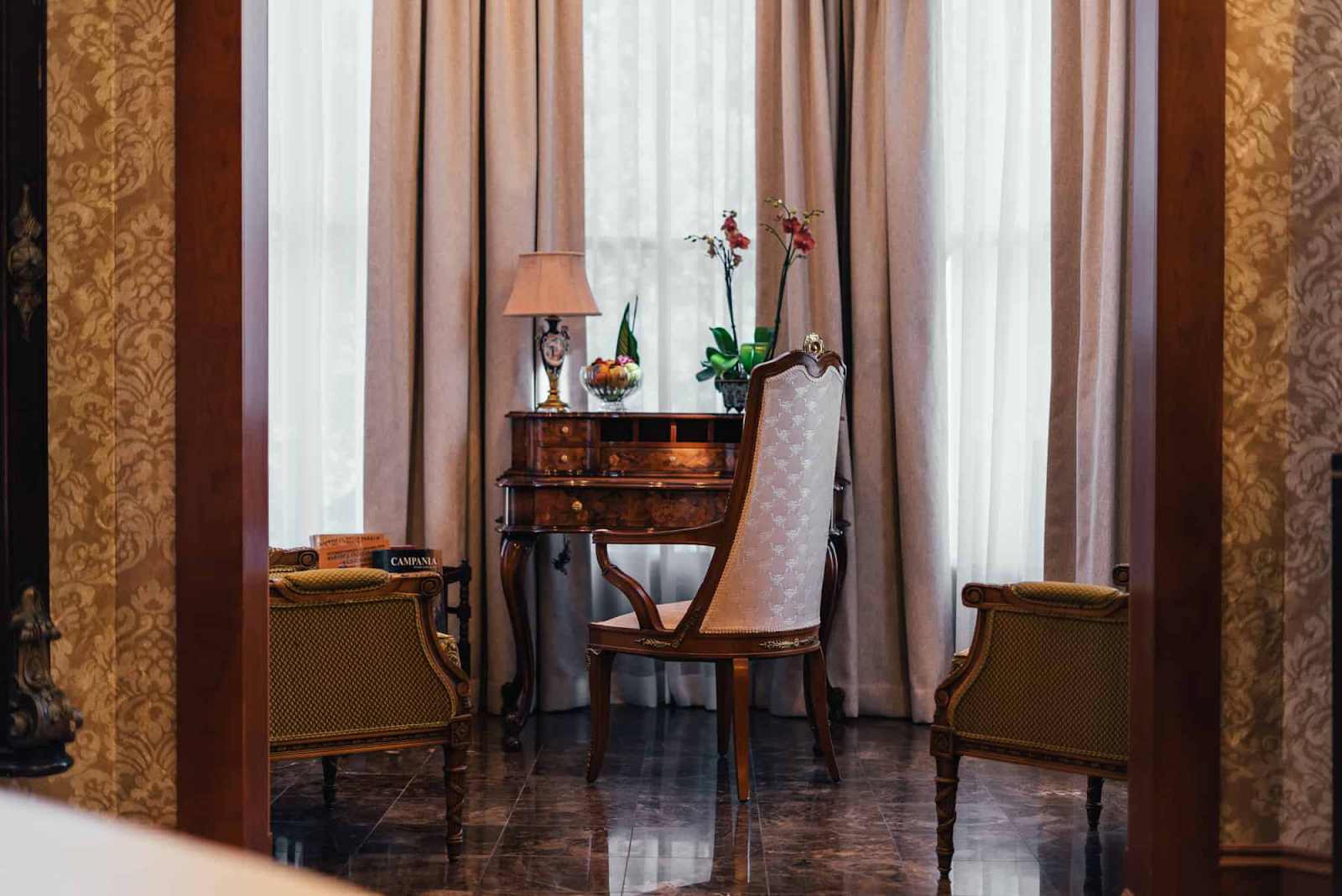 Elegantly furnished room with antique writing desk, orchid and sheer curtains, professionally edited real estate interior photo