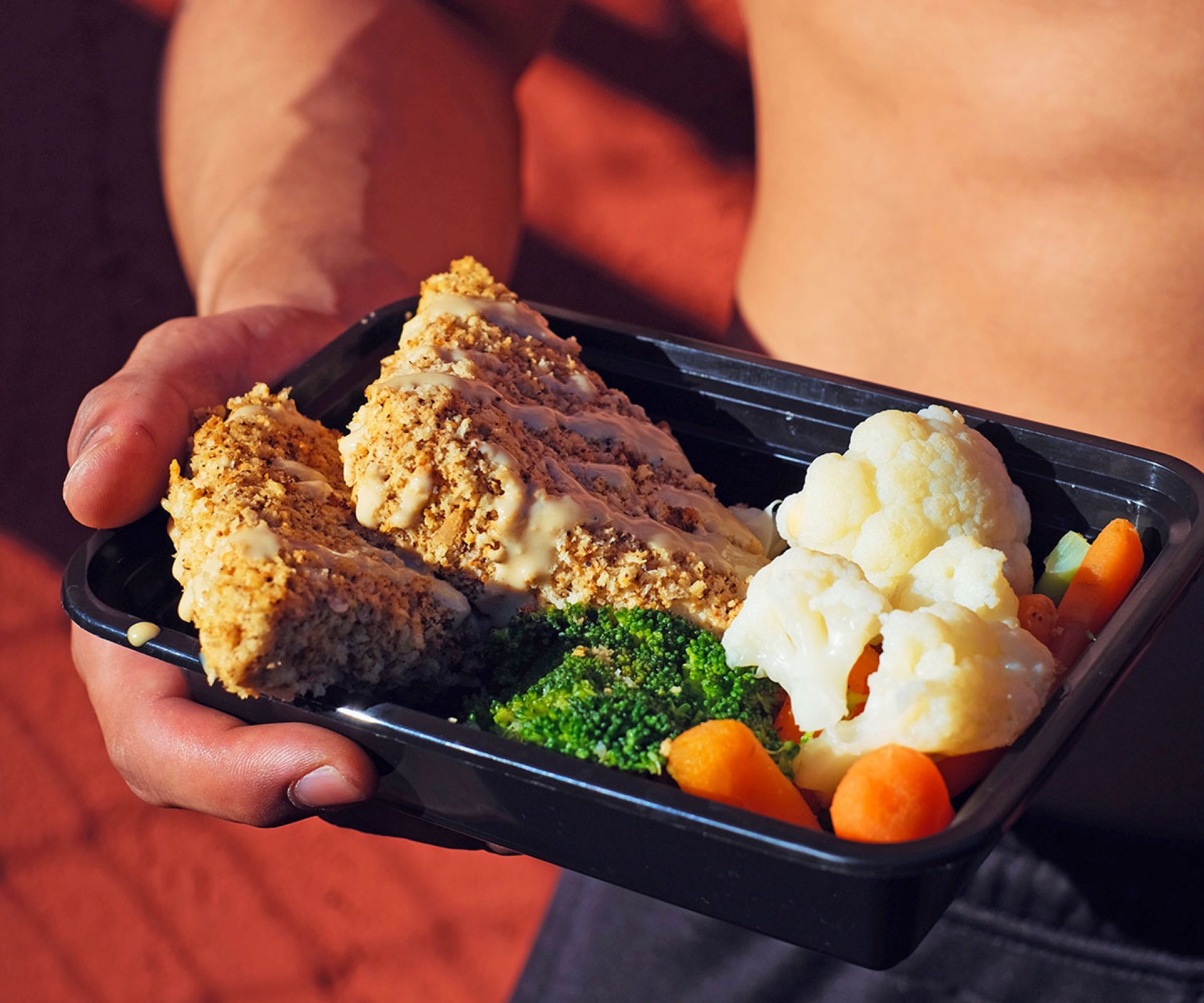 Man holding a meal prep container with crusted chicken, broccoli, carrots, and cauliflower — high-quality food photography to boost healthy restaurant orders