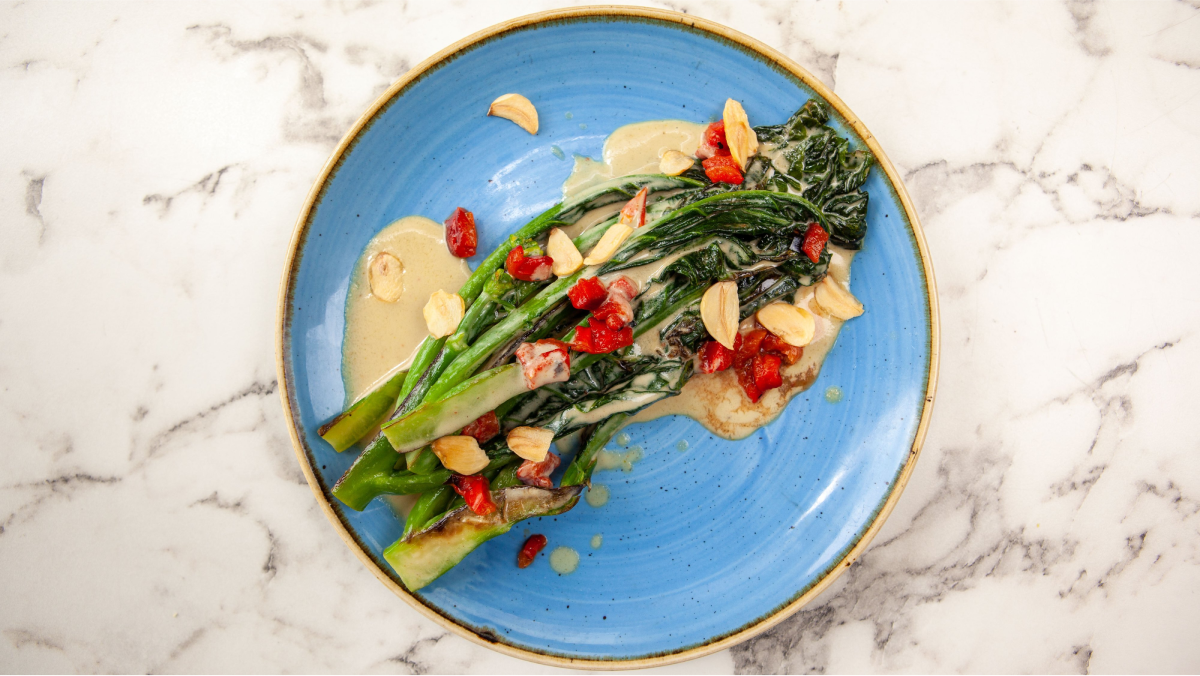 Overhead view of grilled broccolini with tahini sauce, roasted red peppers, and toasted almonds on a blue ceramic plate — Snappr food photography May 2021