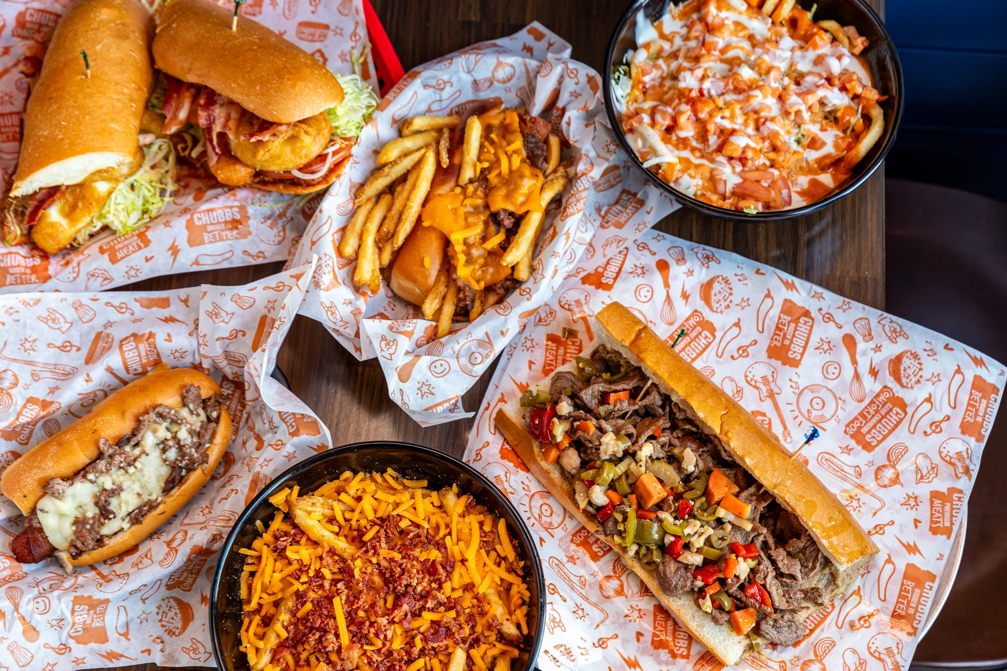 Food photography tip: Flat-lay for showcasing a comfort food spread of loaded steak subs, cheesy fries, and a hot dog on orange wax paper.