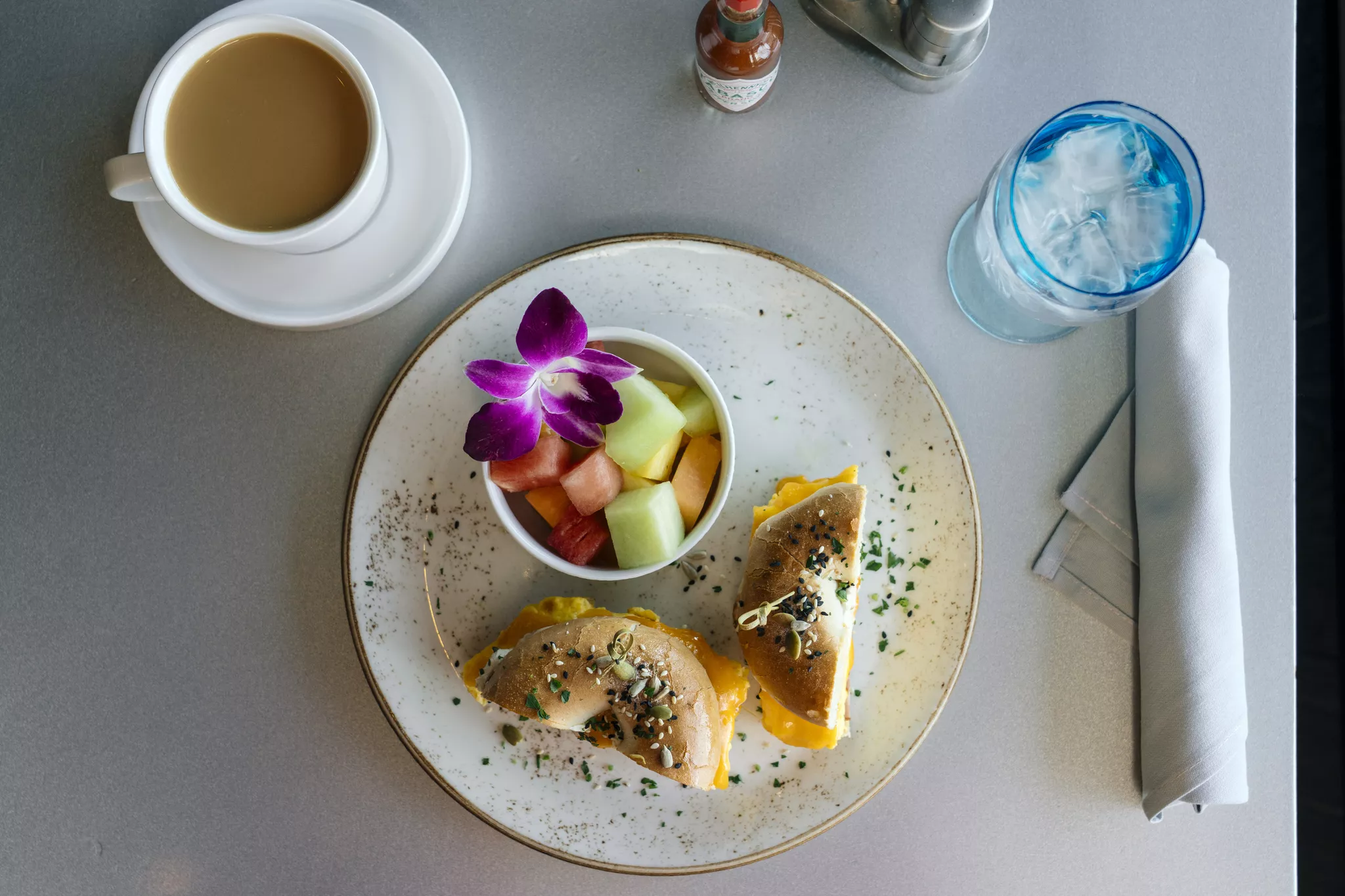 High-angle shot illustrating food photography tips: a bagel sandwich, fresh fruit bowl with a flower, coffee, and iced water on a clean table.