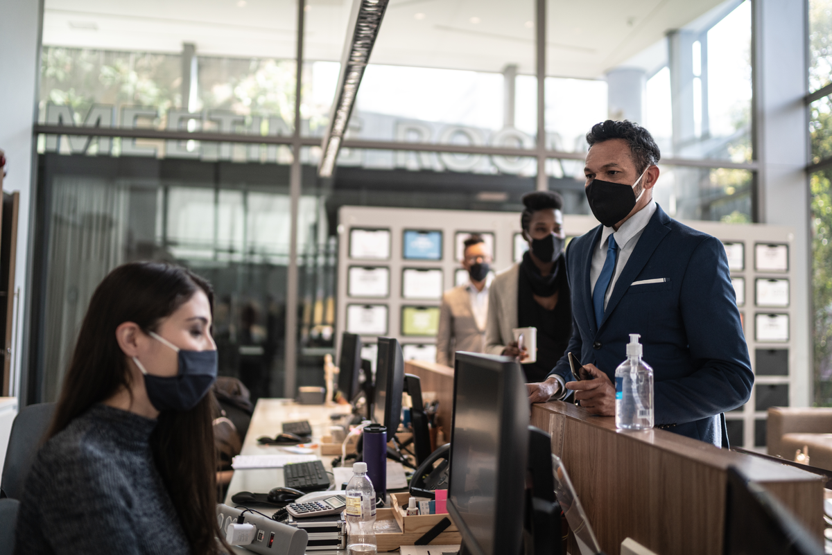 Masked hotel front desk staff checking in a business traveler, illustrating safety protocols businesses adopt to win over travelers during the vacation boom