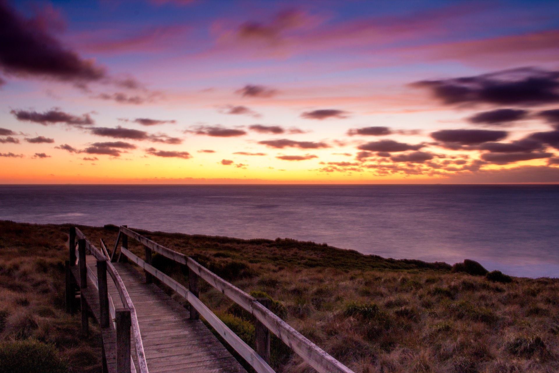 Wooden coastal boardwalk leading toward the ocean at a vibrant purple-orange sunset, near Quat Quatta one of Melbourne's most scenic wedding venues