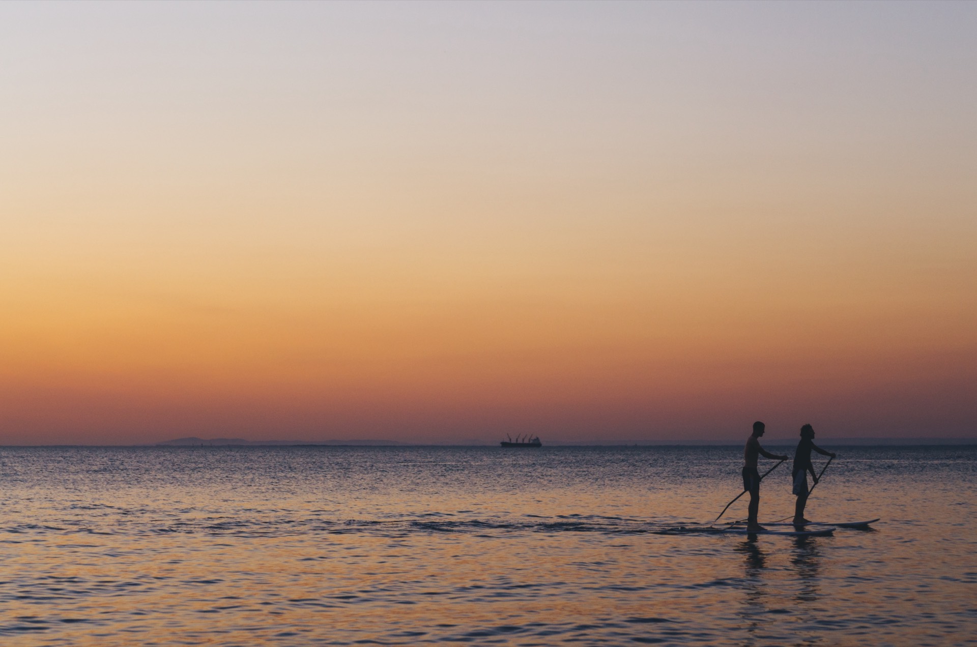 Two silhouetted paddleboarders on calm ocean water at a golden-orange sunset, evoking the romantic outdoor ambience of Stones of the Yarra Valley wedding venue