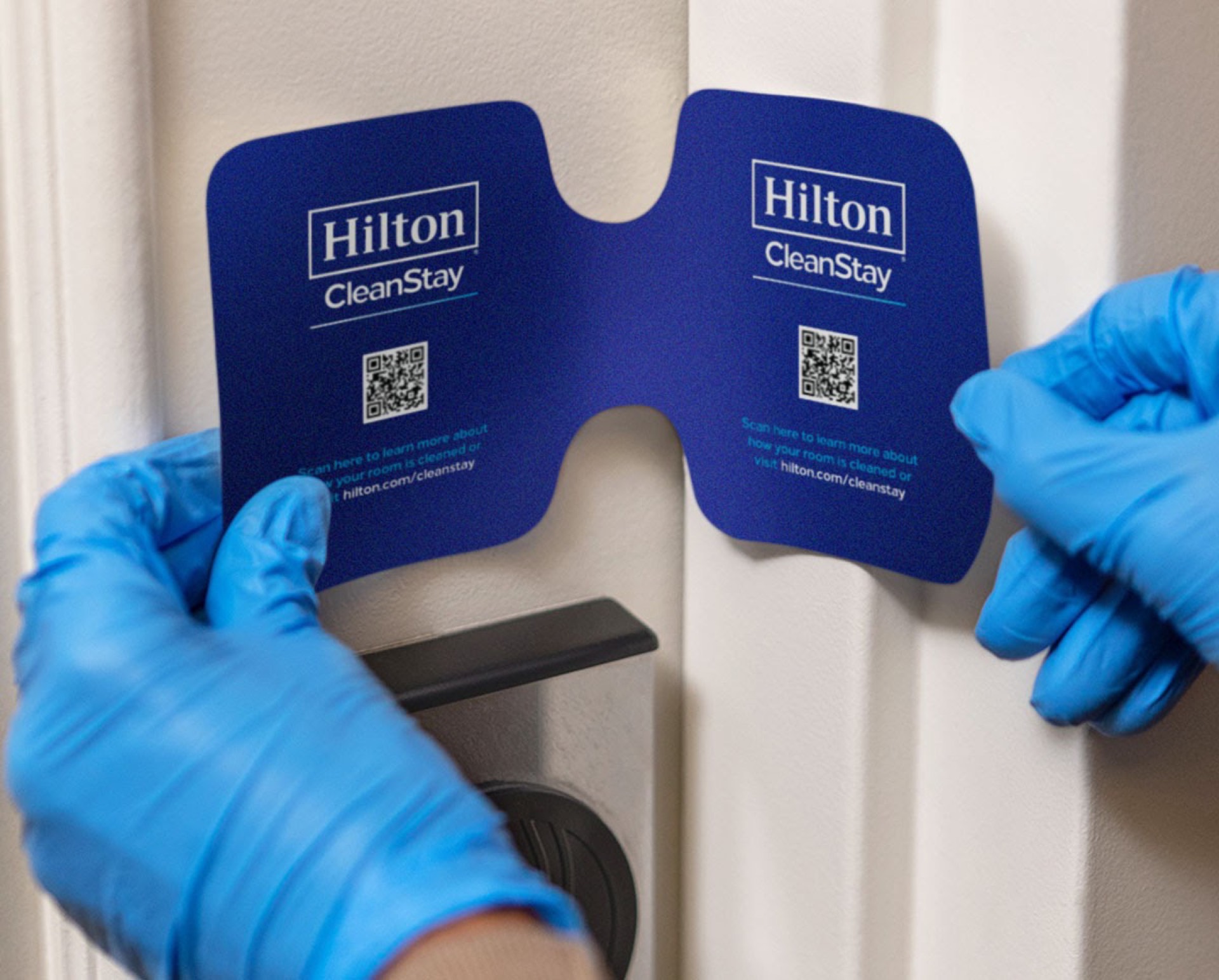 Hilton CleanStay seal being placed on a hotel room door handle by gloved hands, highlighting COVID cleanliness protocols