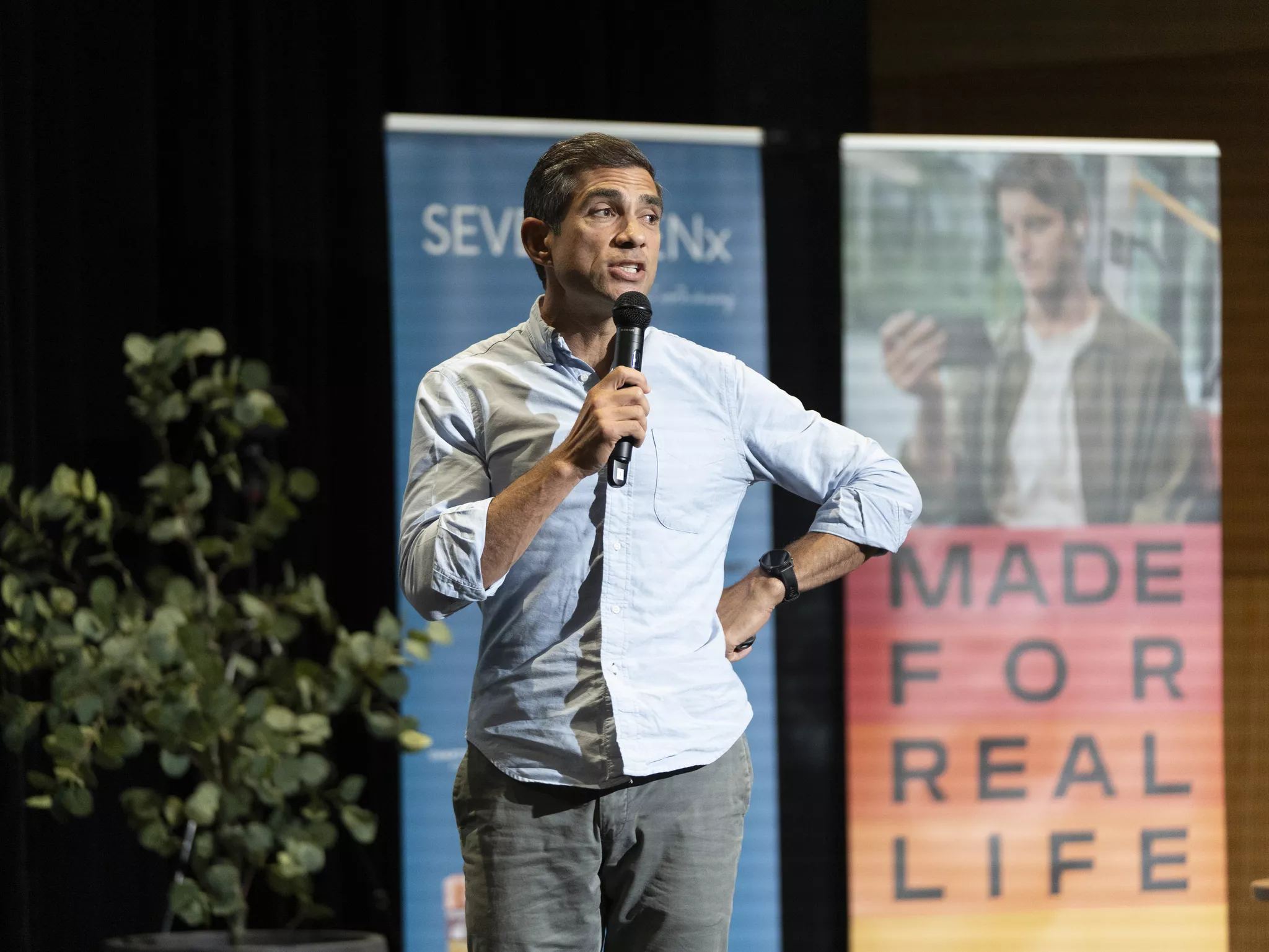 Speaker on stage holding a microphone at a corporate event with branded banners behind.