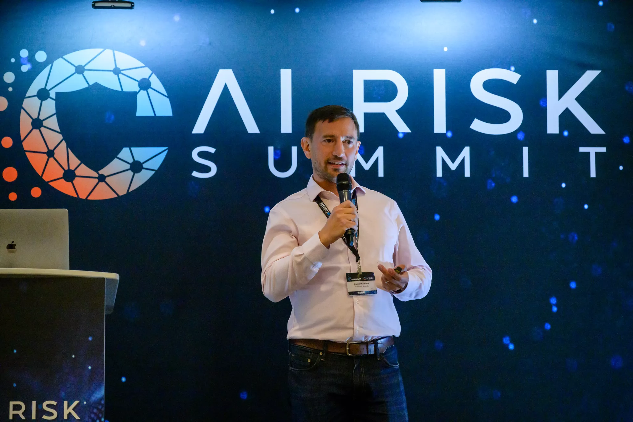 Male speaker with microphone on stage before AI Risk Summit branded backdrop .