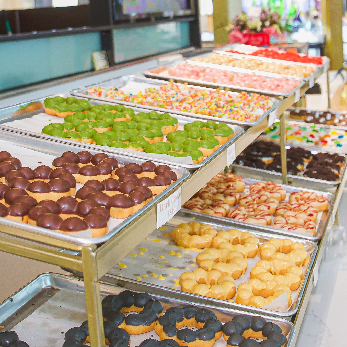 Colorful bakery display with trays of assorted donuts including chocolate, green tea, and sprinkle varieties