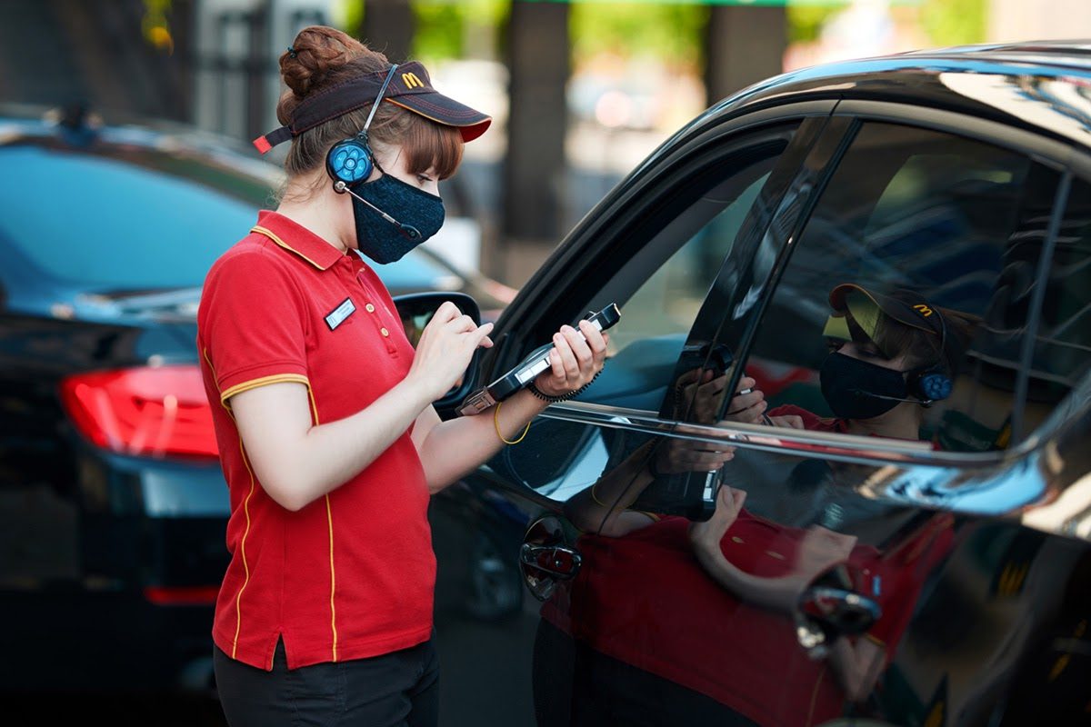 McDonald's employee with headset and face mask taking drive-thru order at car window using handheld device