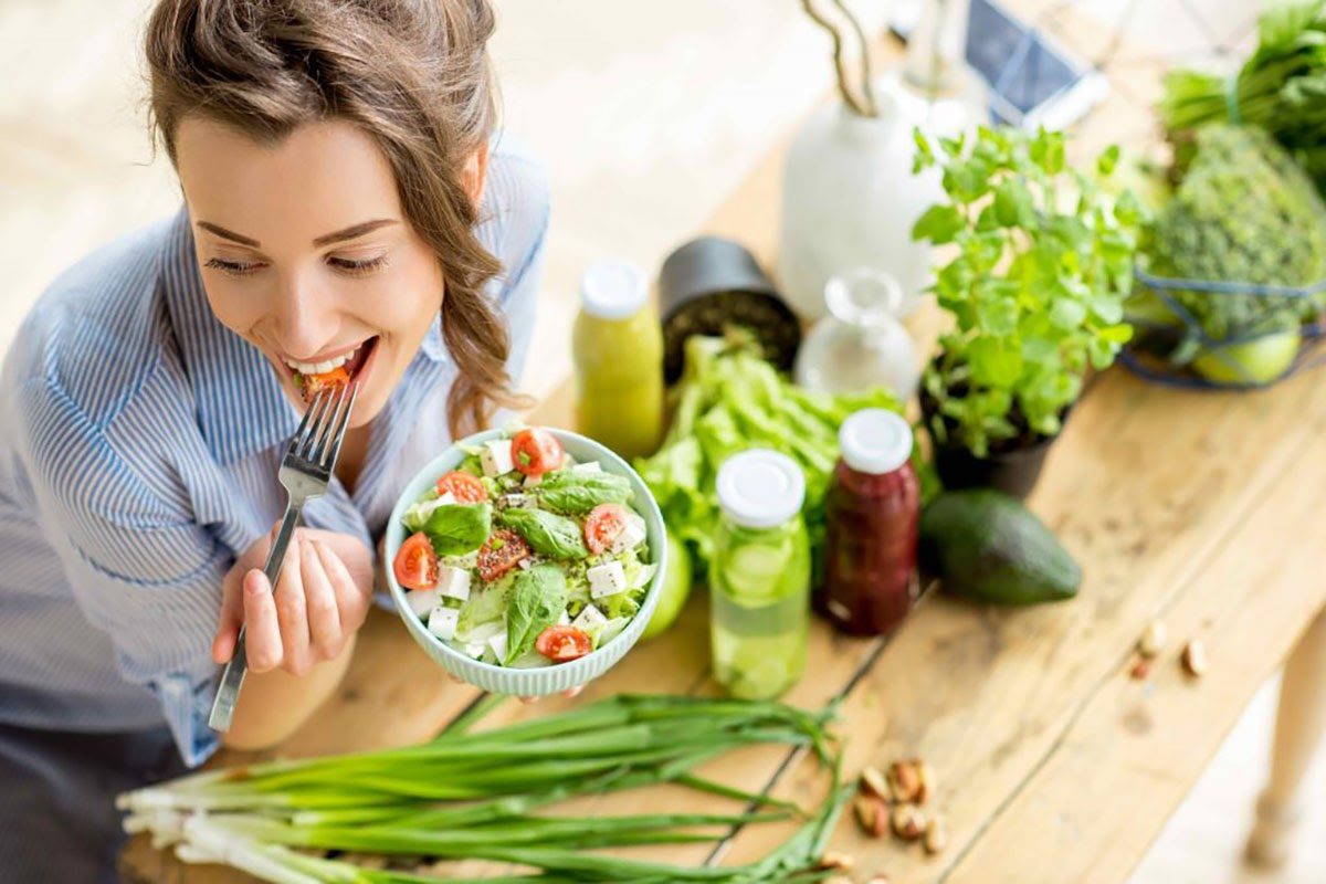 Woman eating a fresh green salad with tomatoes and feta surrounded by vegetables and juice bottles