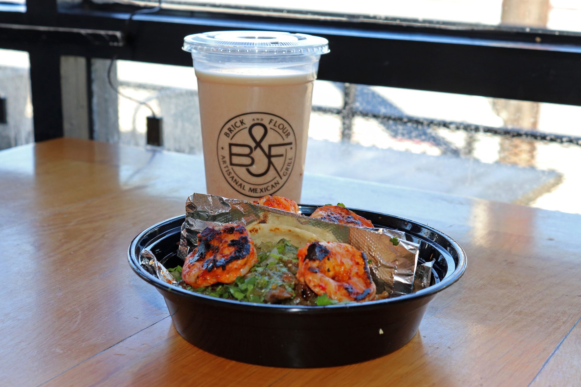 Grilled shrimp bowl with salsa verde in a takeout container alongside a branded cup at a restaurant patio
