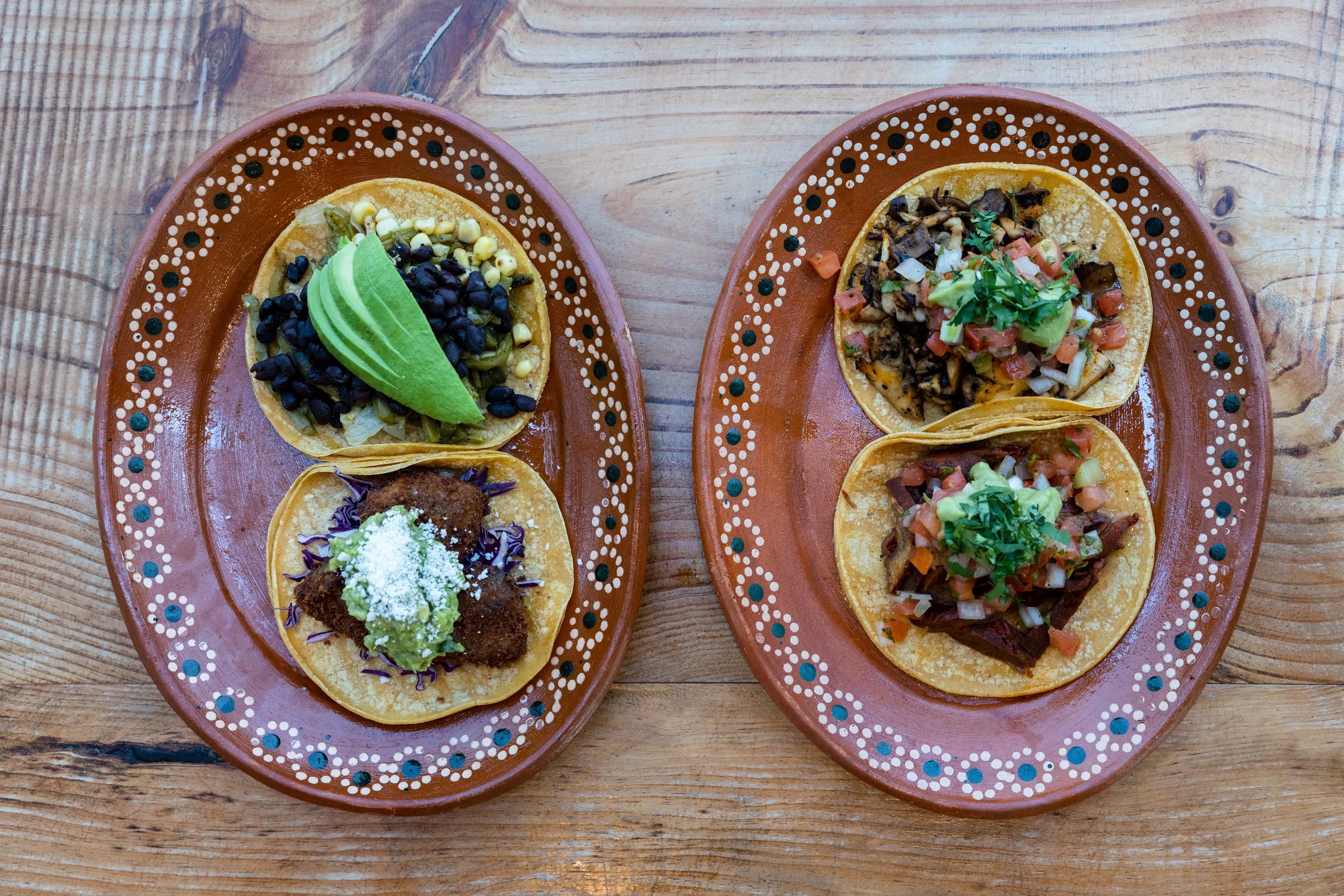 Overhead view of four colorful tacos with fresh toppings on decorative Mexican clay plates on a wooden table