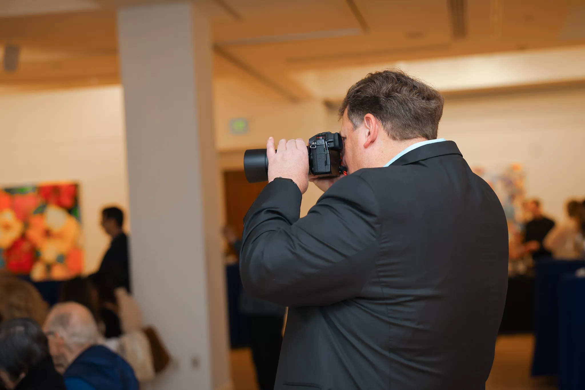 Event photographer in a suit shooting guests at an indoor gallery reception.