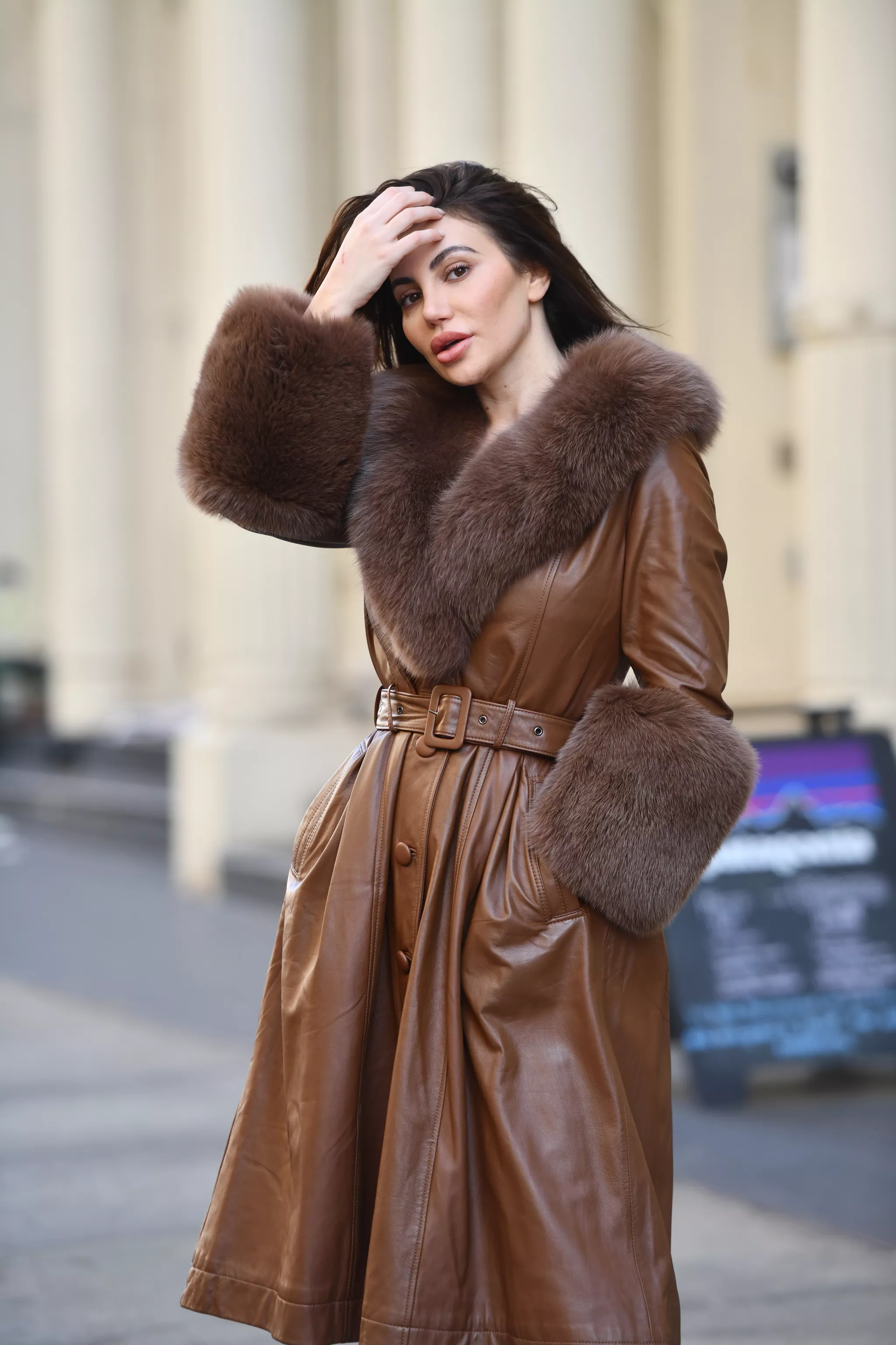 Fashion editorial of a woman in a brown leather coat with fur collar on a street.