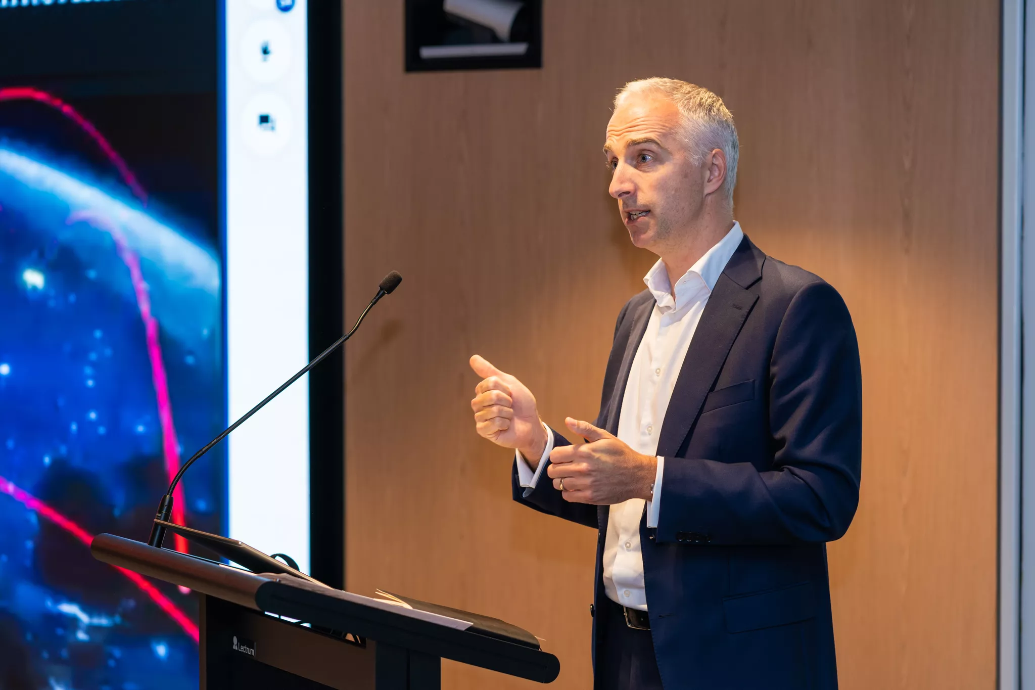 Event editorial photo of a speaker presenting at a lectern beside a screen.