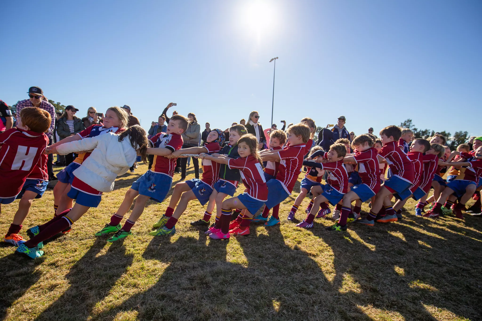 Editorial event photo of children in team kit playing tug-of-war in backlit sun.