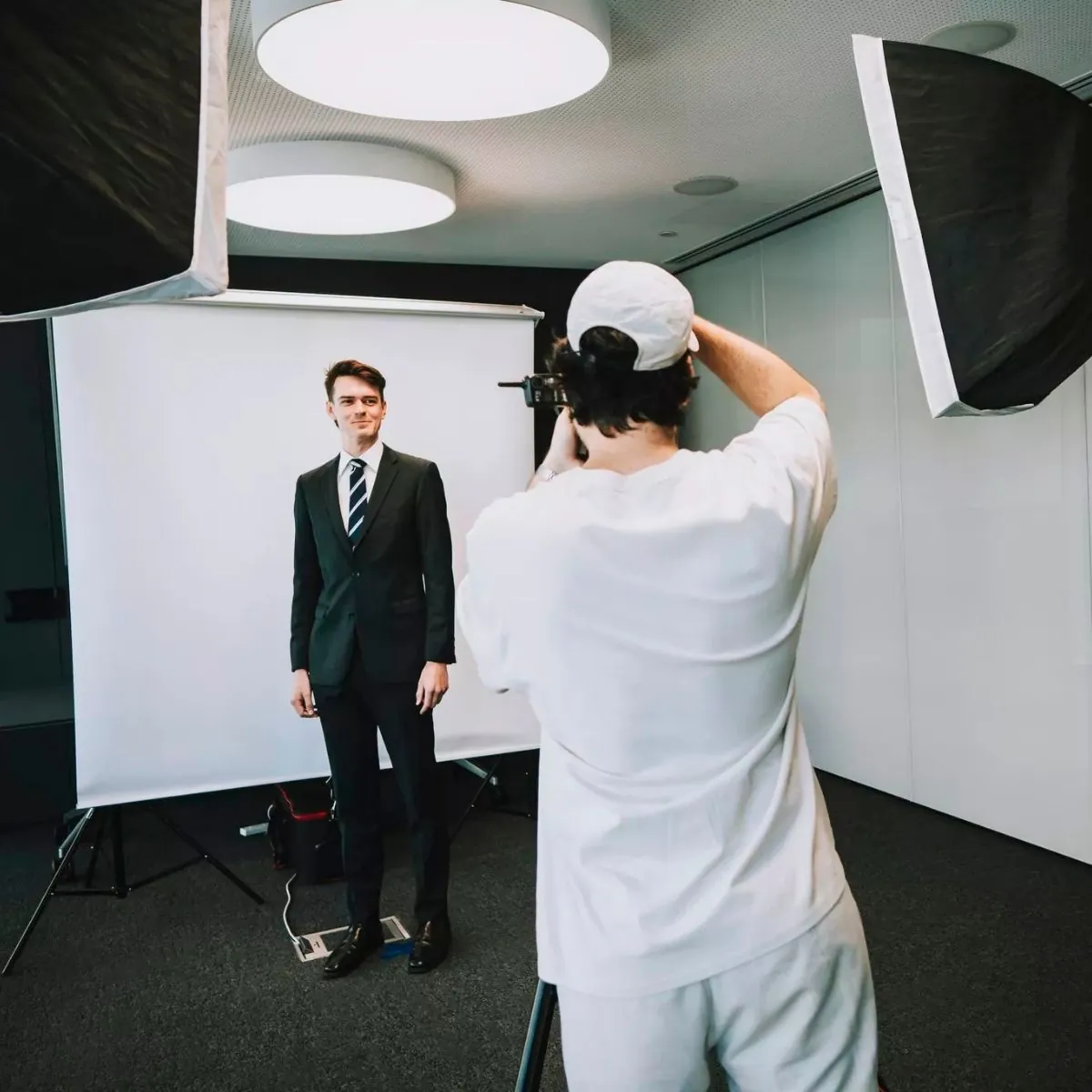 Photographer taking a professional headshot of a man in a dark suit and striped tie, standing in front of a white backdrop with studio lighting