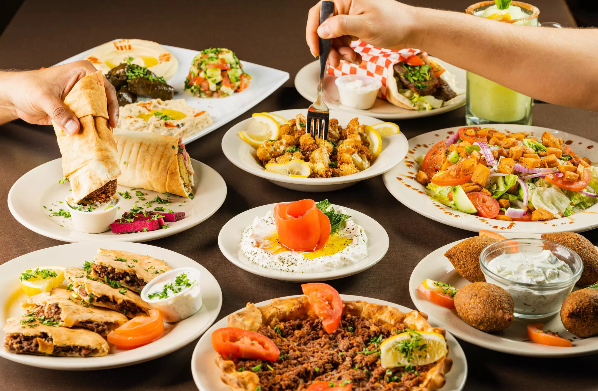 Hands reaching across a mezze spread of hummus, falafel and kibbeh styled for a restaurant menu.