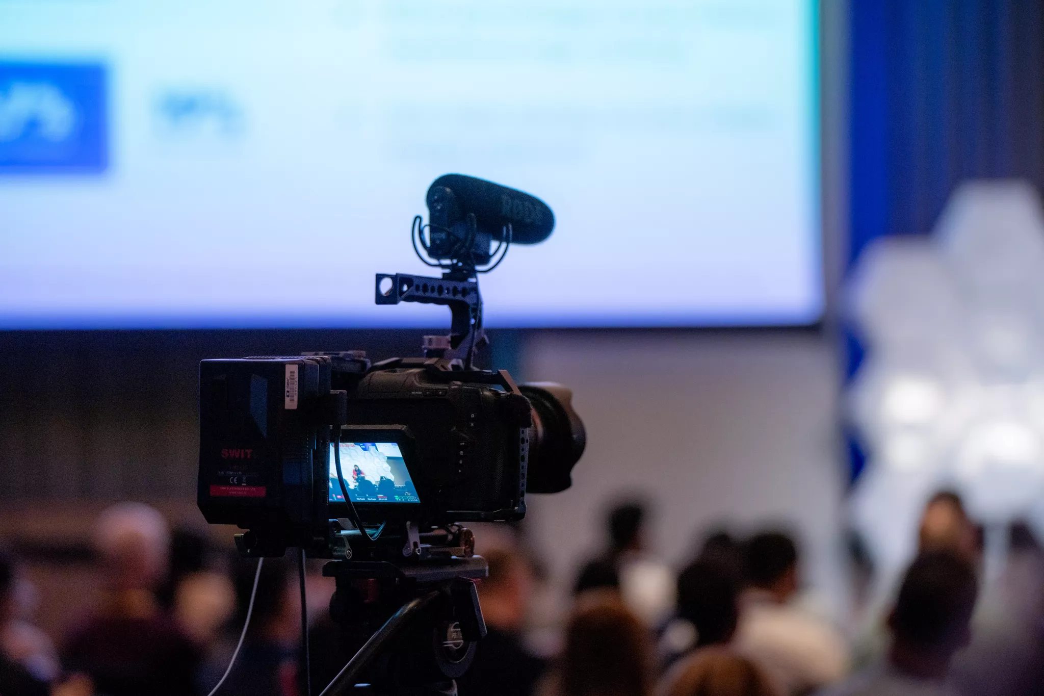 Video production service camera and shotgun mic trained on a conference stage screen.
