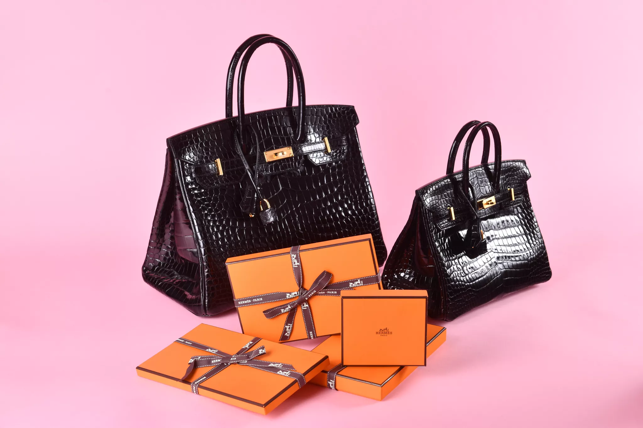 Group product photography of two crocodile handbags and orange gift boxes on pink.
