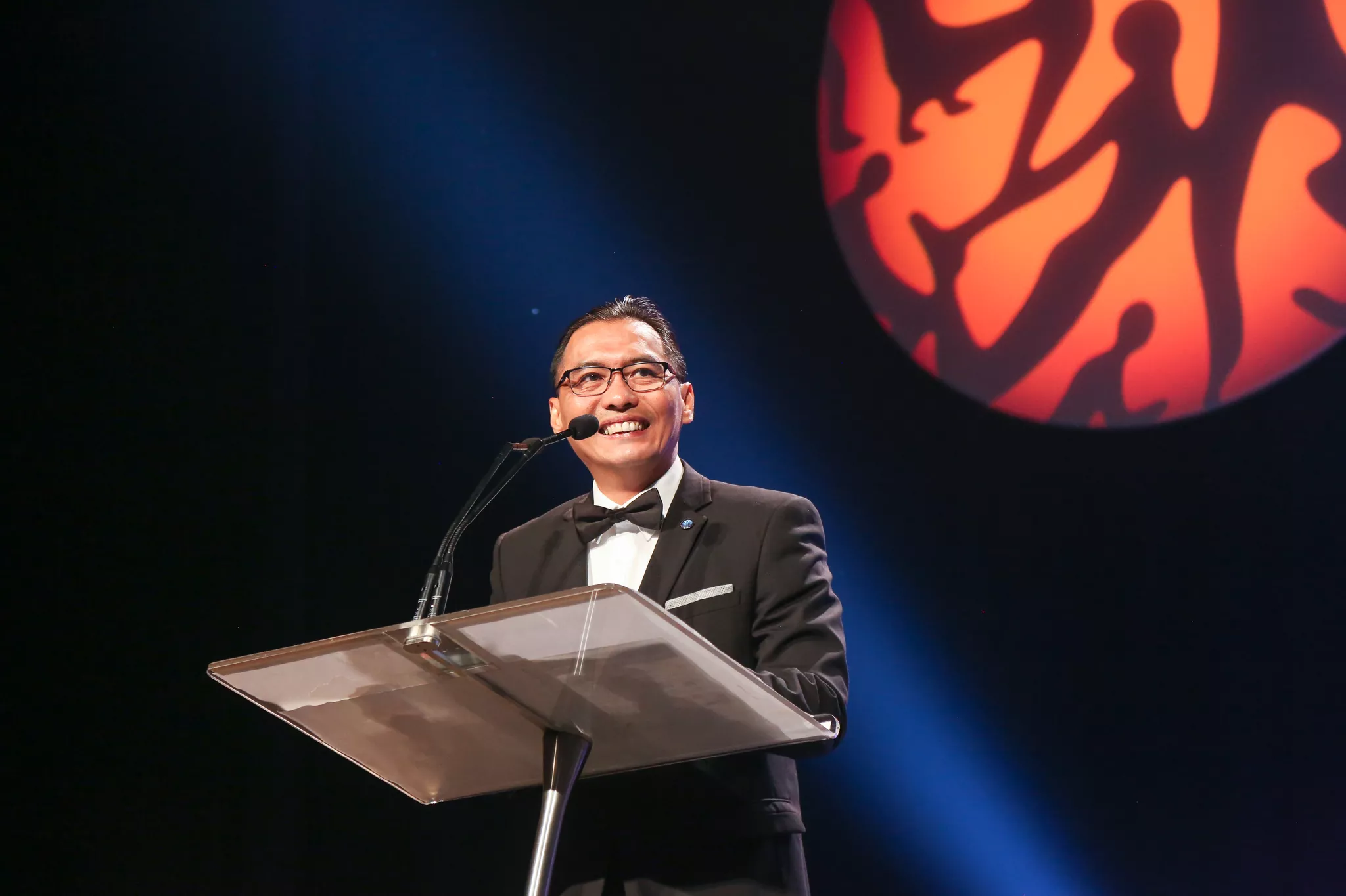 Keynote speaker at a lectern under stage lighting, shot by a convention photographer.