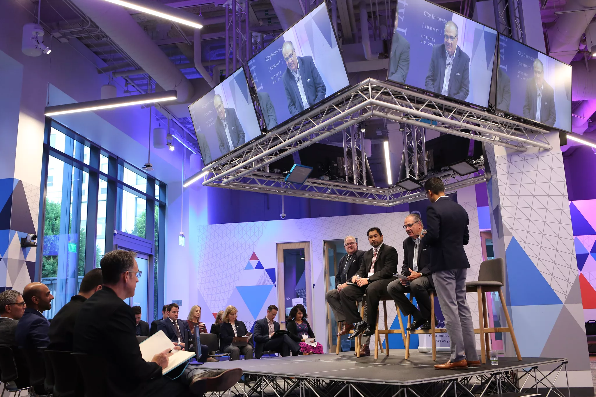 Panel discussion on a summit stage under overhead screens, shot by a convention photographer.