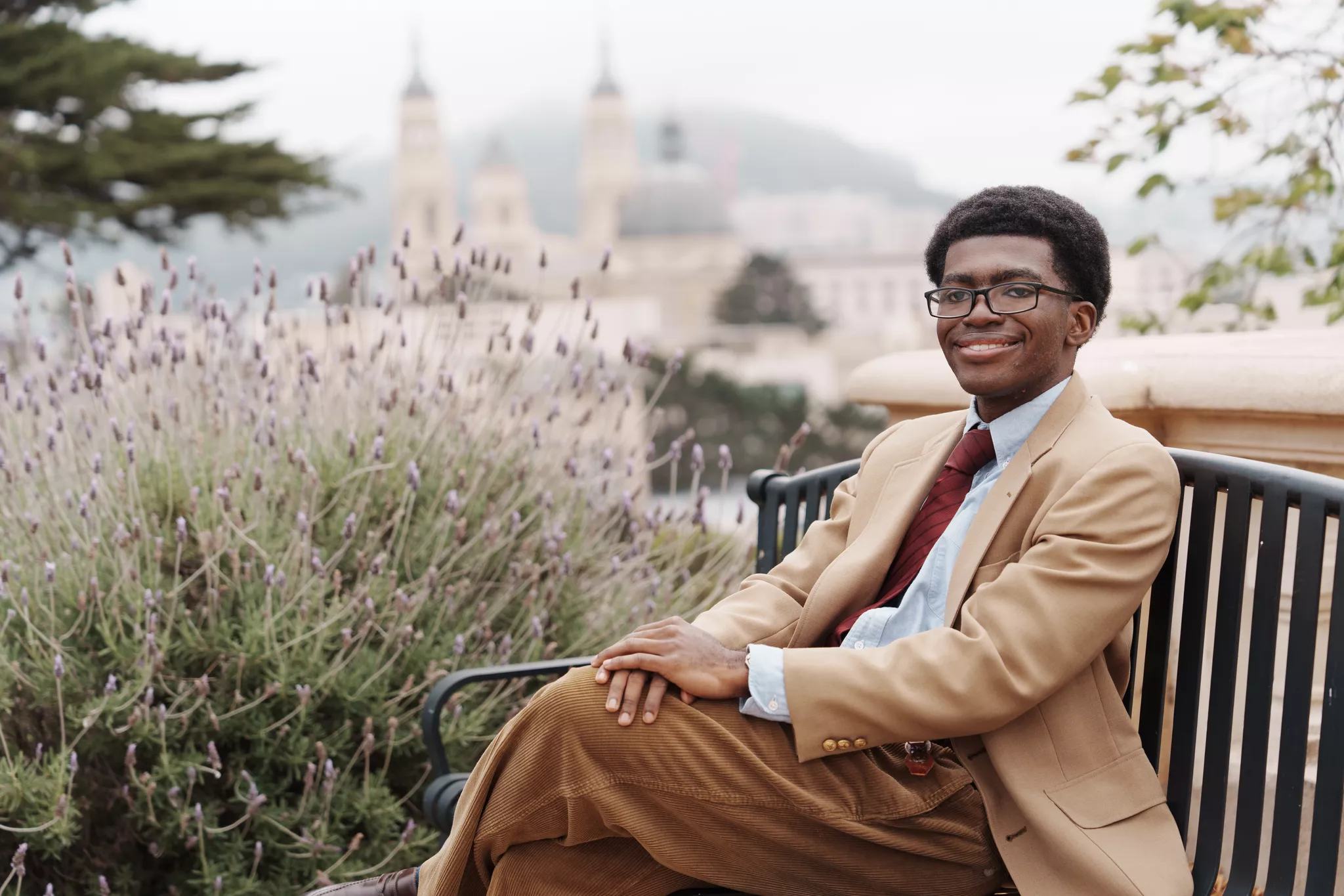 Young man in a tan blazer and tie seated on a bench, legs crossed, fog behind him