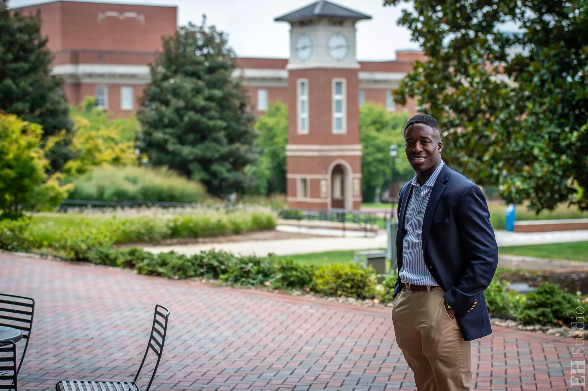 Young man in a navy blazer and khakis, hands in pockets, on a brick campus plaza