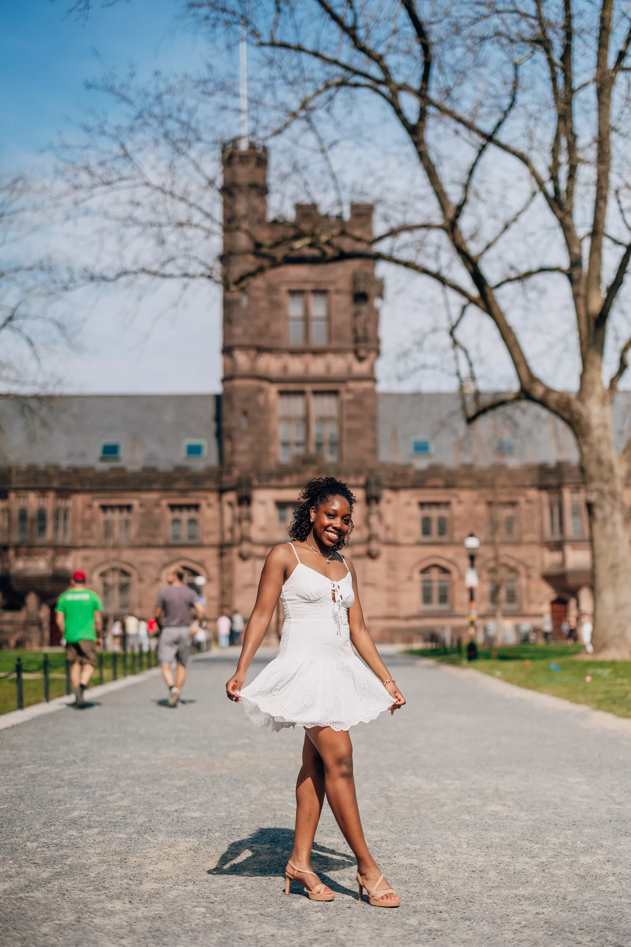 Young woman in a white sundress holding her skirt out, campus tower behind her.
