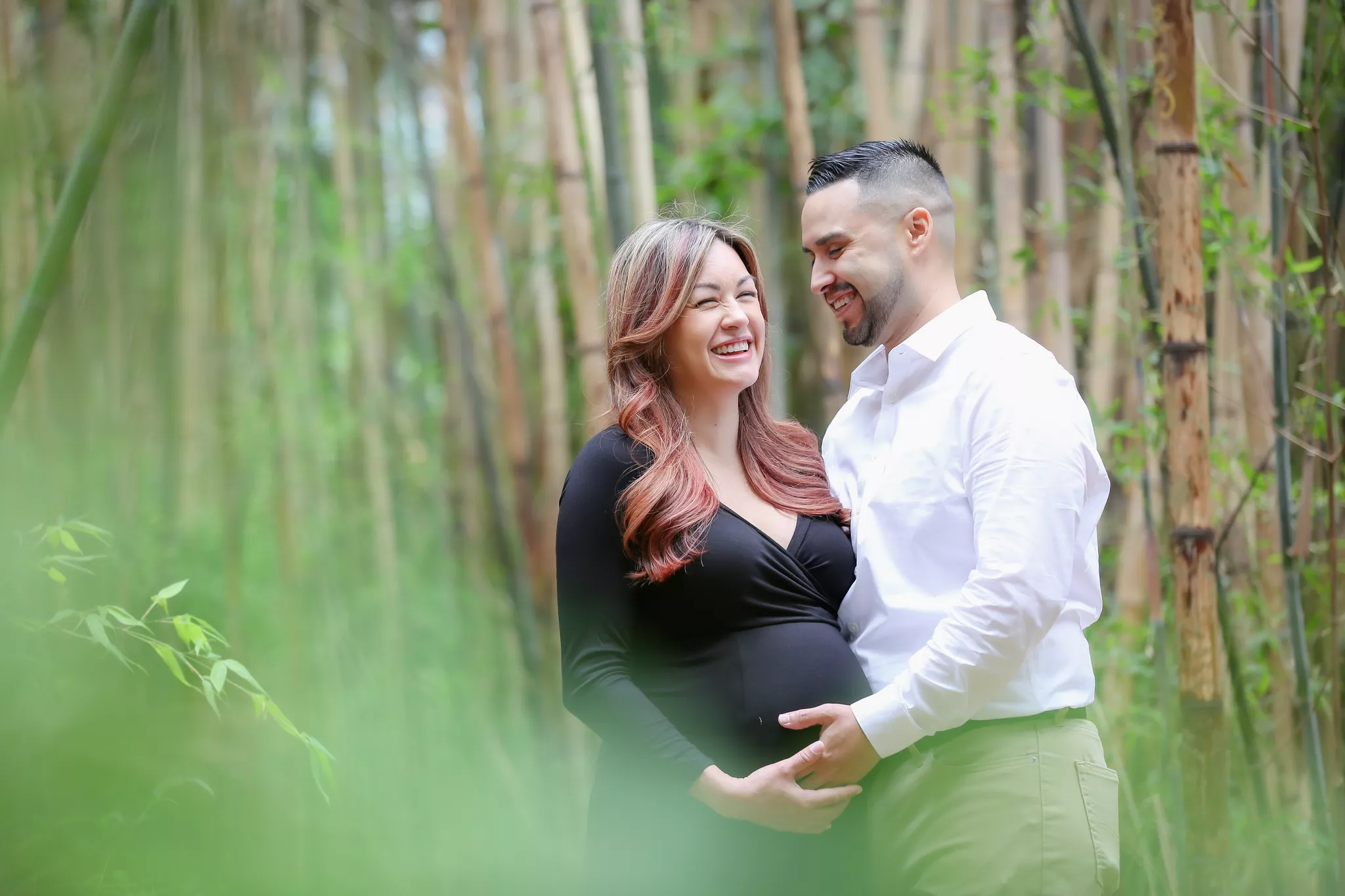 Maternity photo poses with a partner — laughing couple, both hands cradling the bump in bamboo.