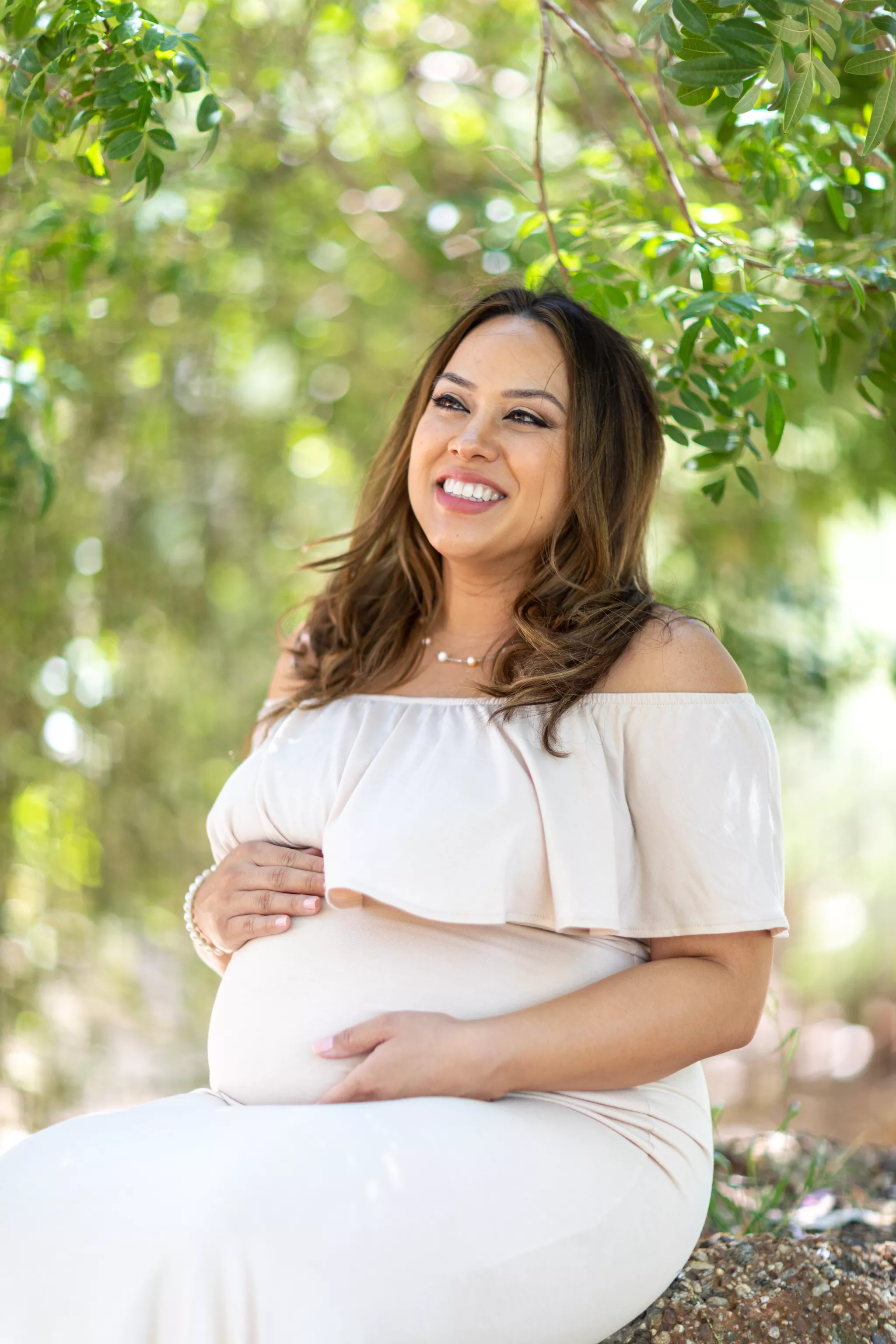 Maternity photo poses seated under a tree — one hand under the bump, one resting above