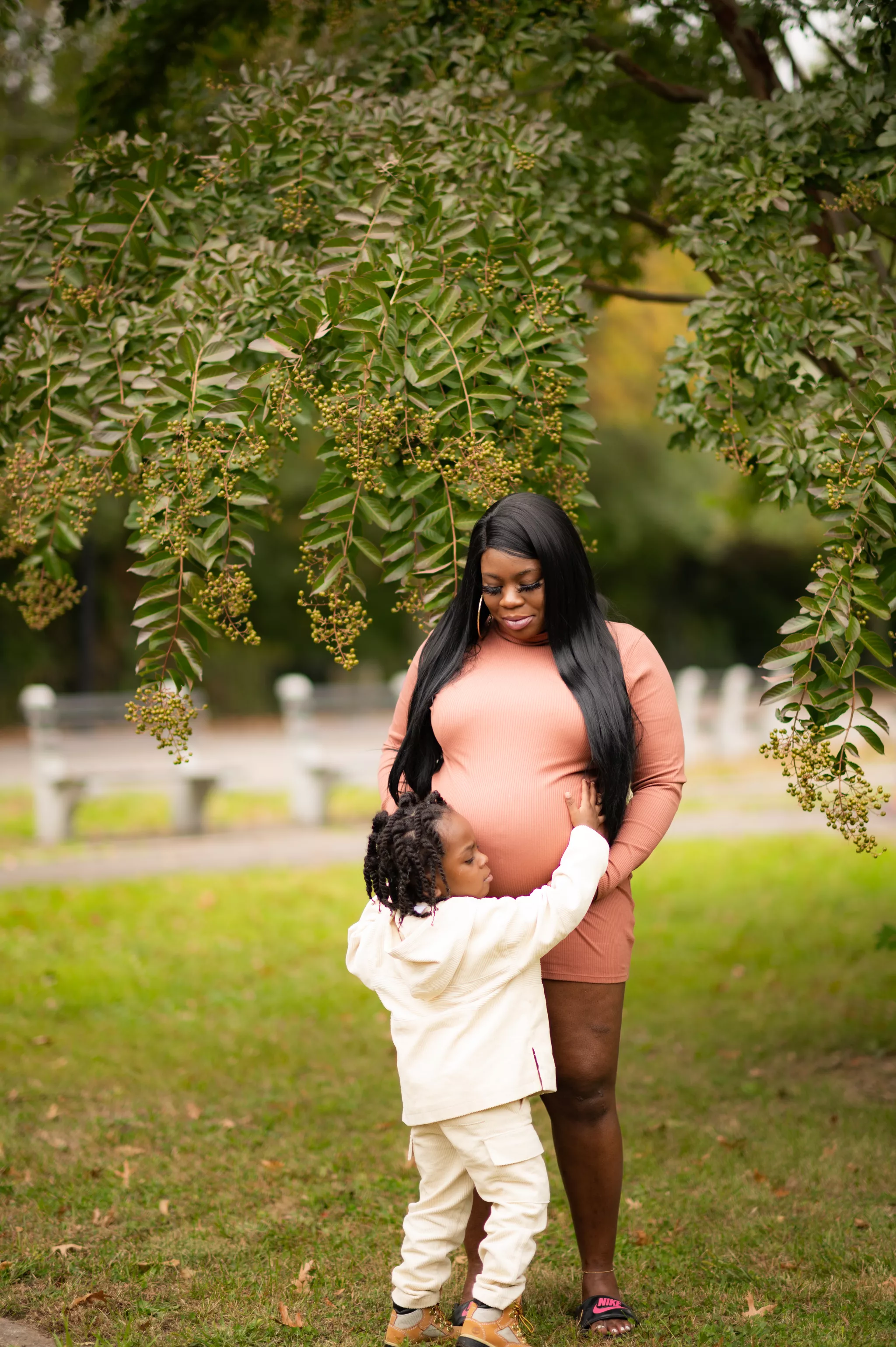 Maternity photo poses with a child — young child leans in to kiss the bump under a park tree